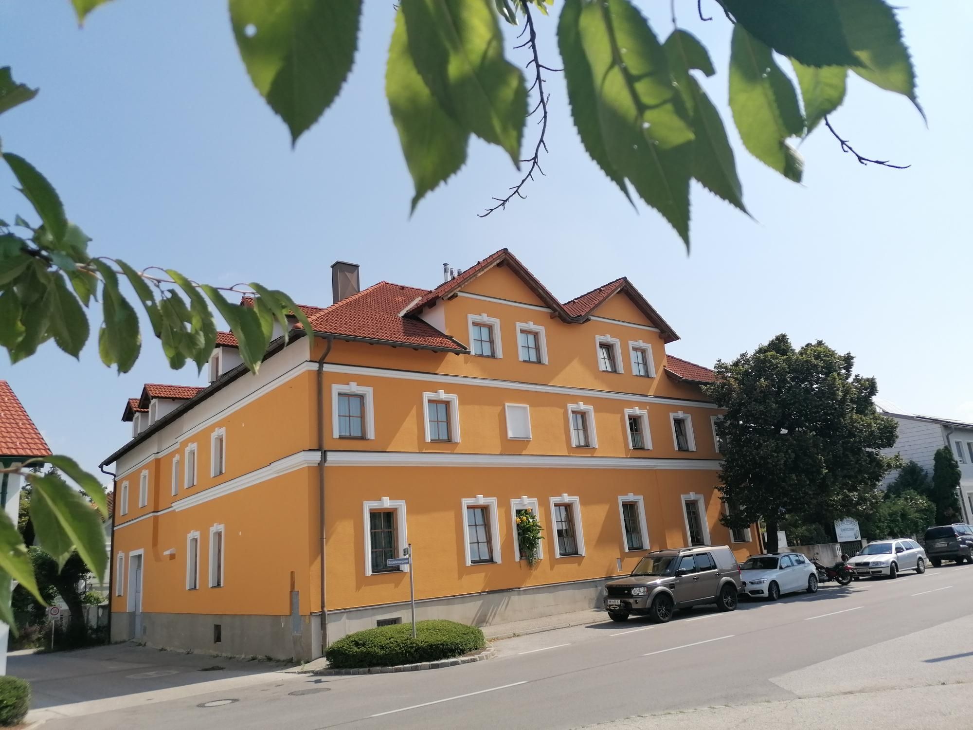 Žltá budova s červenými strechami, obklopená stromami a zaparkovanými autami.