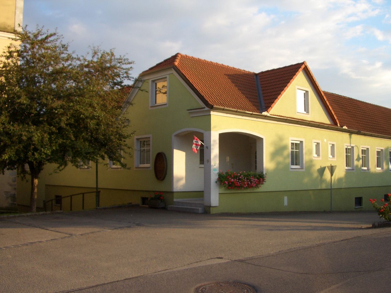 Žltá budova s červenou strechou a kvetmi pri vchode.