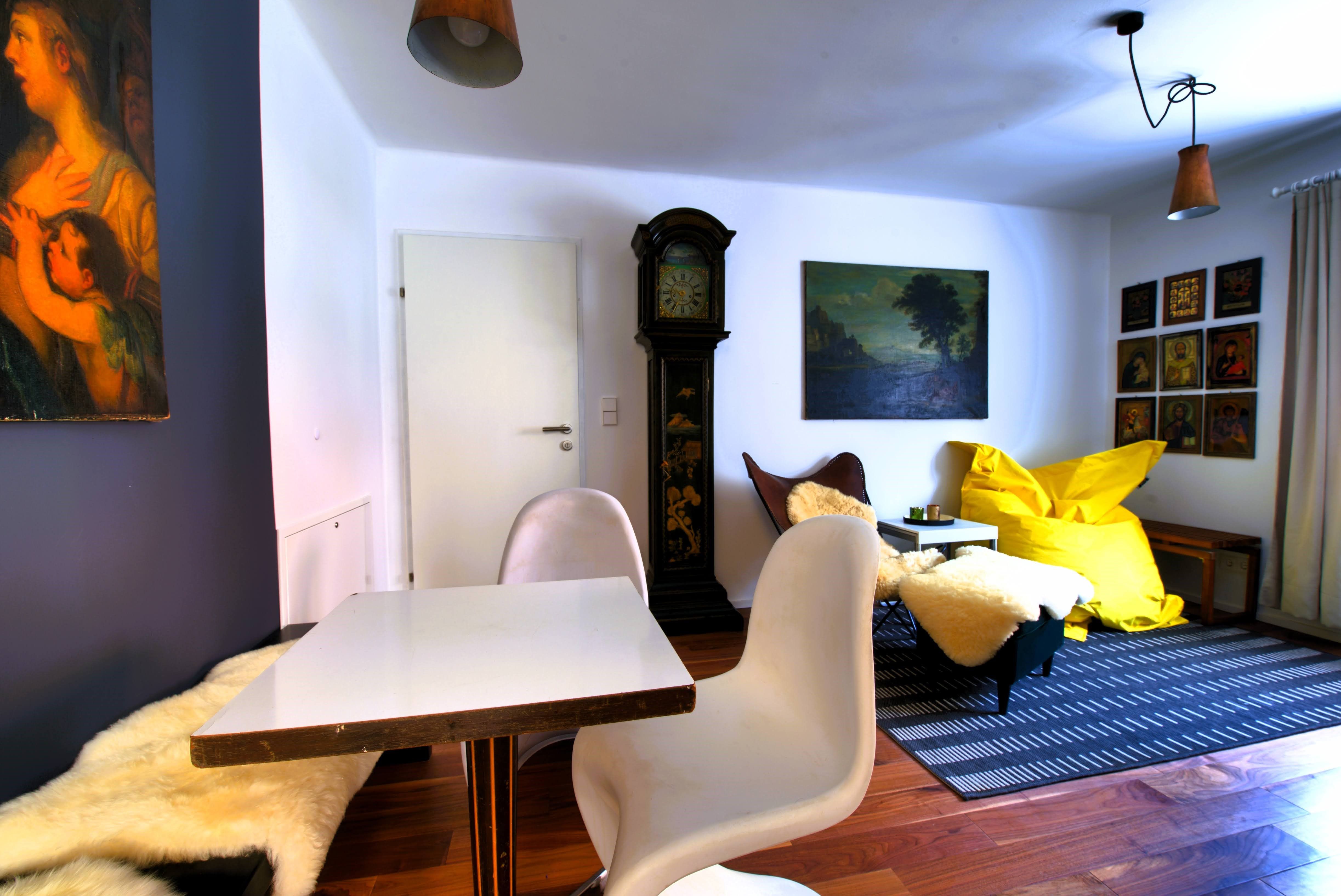 Obývacia izba so stolom, stoličkami, dedkovskými hodinami, obrazmi a žltým fazuľovým vankúšom.