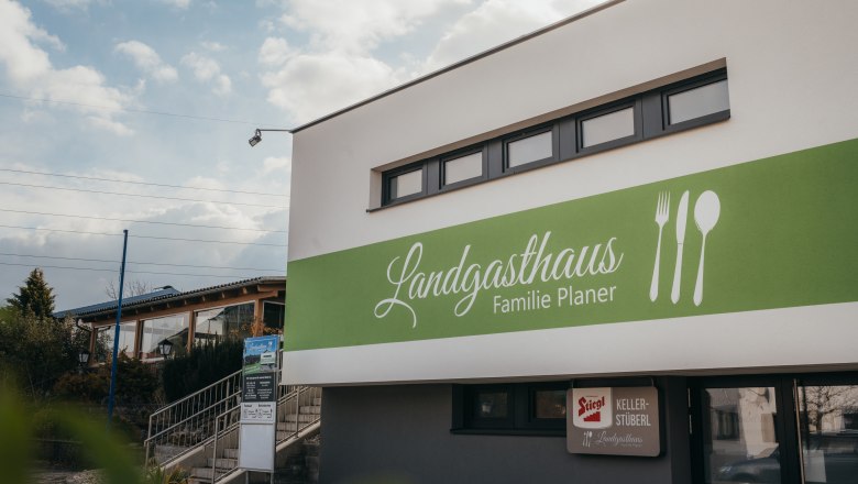 Exteriérový pohľad na vidiecky hostinec rodiny Planerovcov so zeleným nápisom a symbolmi príborov.