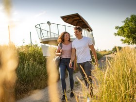 Dvojica kr&aacute;ča po ceste vedľa vyhliadkovej plo&scaron;iny na Wagrame, obklopen&aacute; vysokou tr&aacute;vou a pod jasnou oblohou.