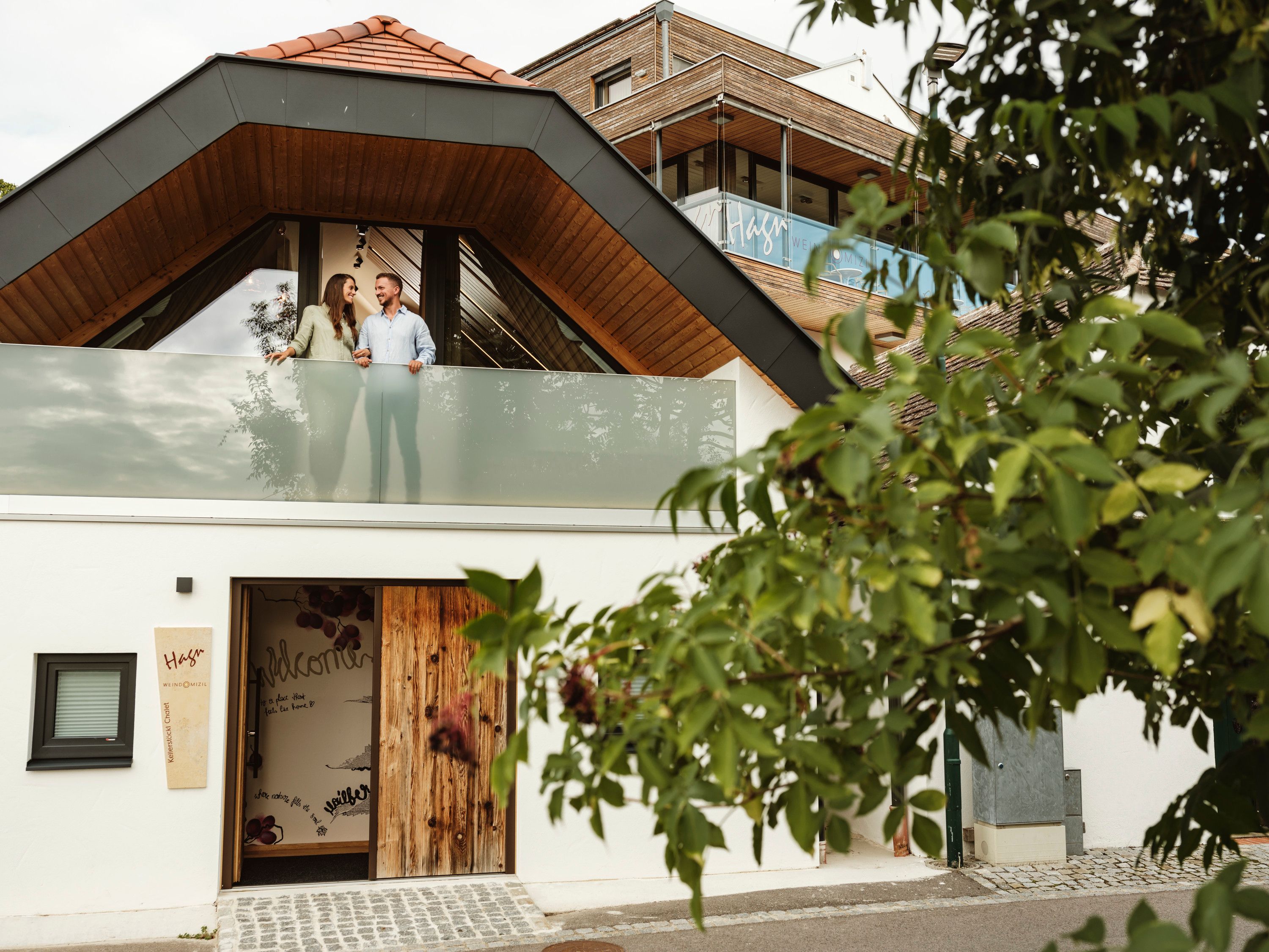 Moderná budova s párom na balkóne, obklopená stromami.