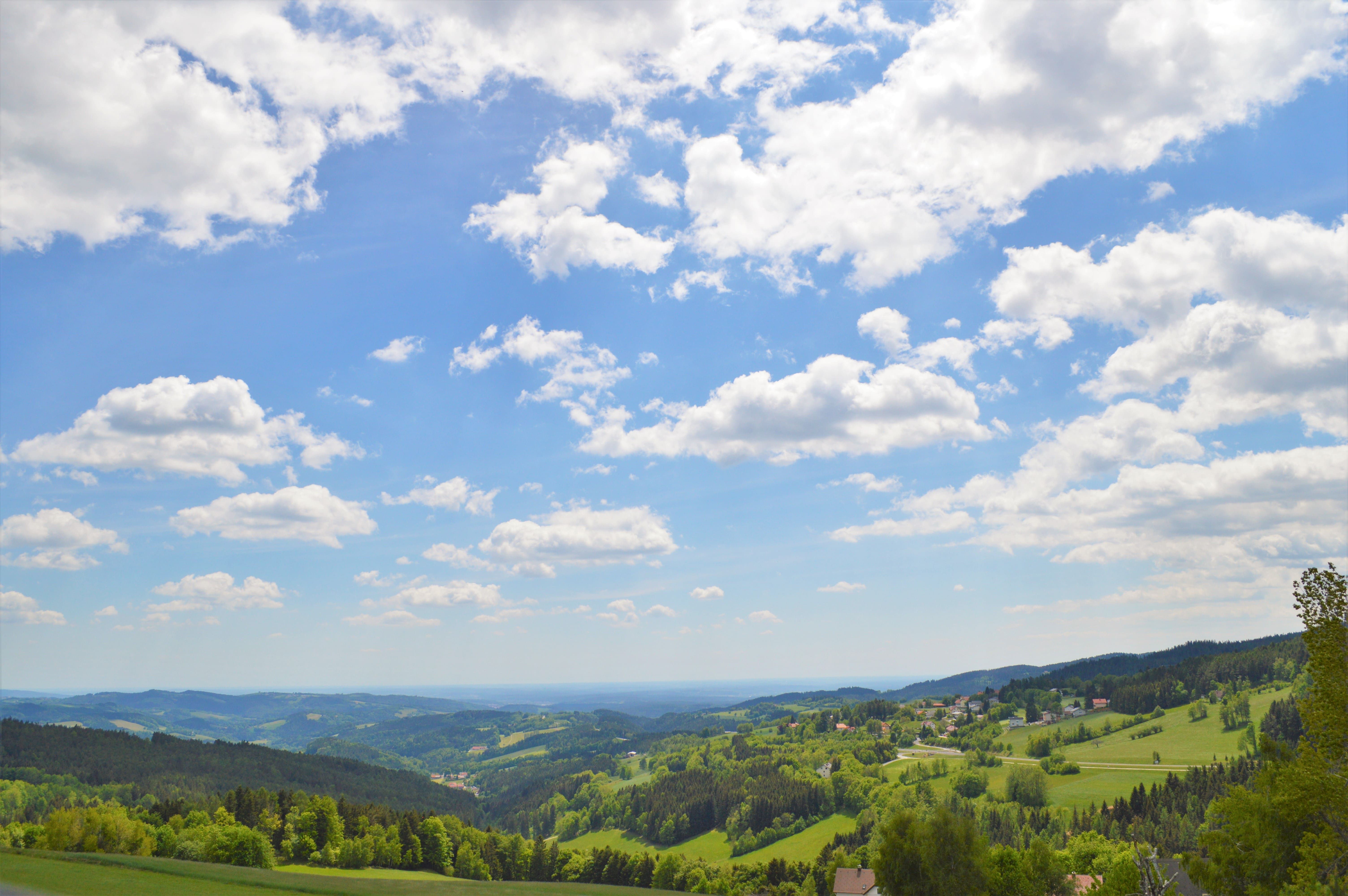 Panoramatický pohľad na kopcovitú krajinu s lesmi a lúkami pod modrou oblohou s bielymi mrakmi.