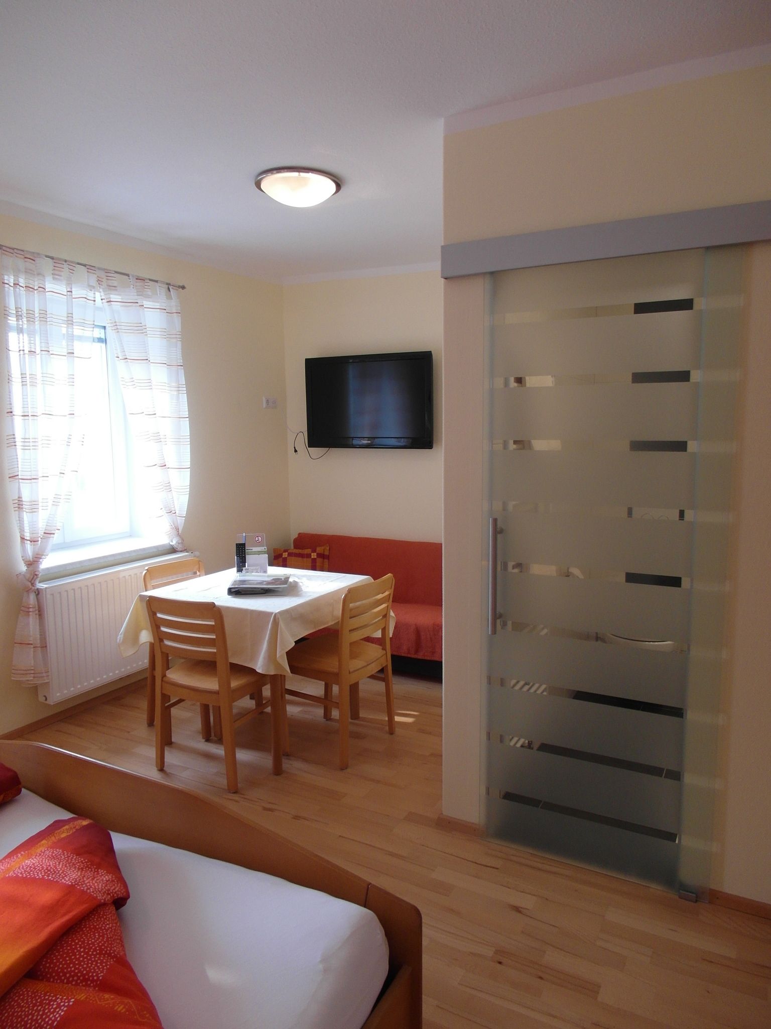 Svetlá izba s jedálenským kútom, stolom, stoličkami, posteľou a TV s plochou obrazovkou.