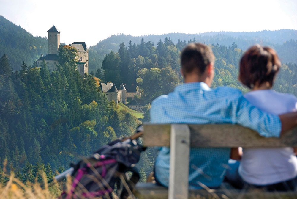Pár sedí na lavičke s výhľadom na hrad v zalesnenej krajine.