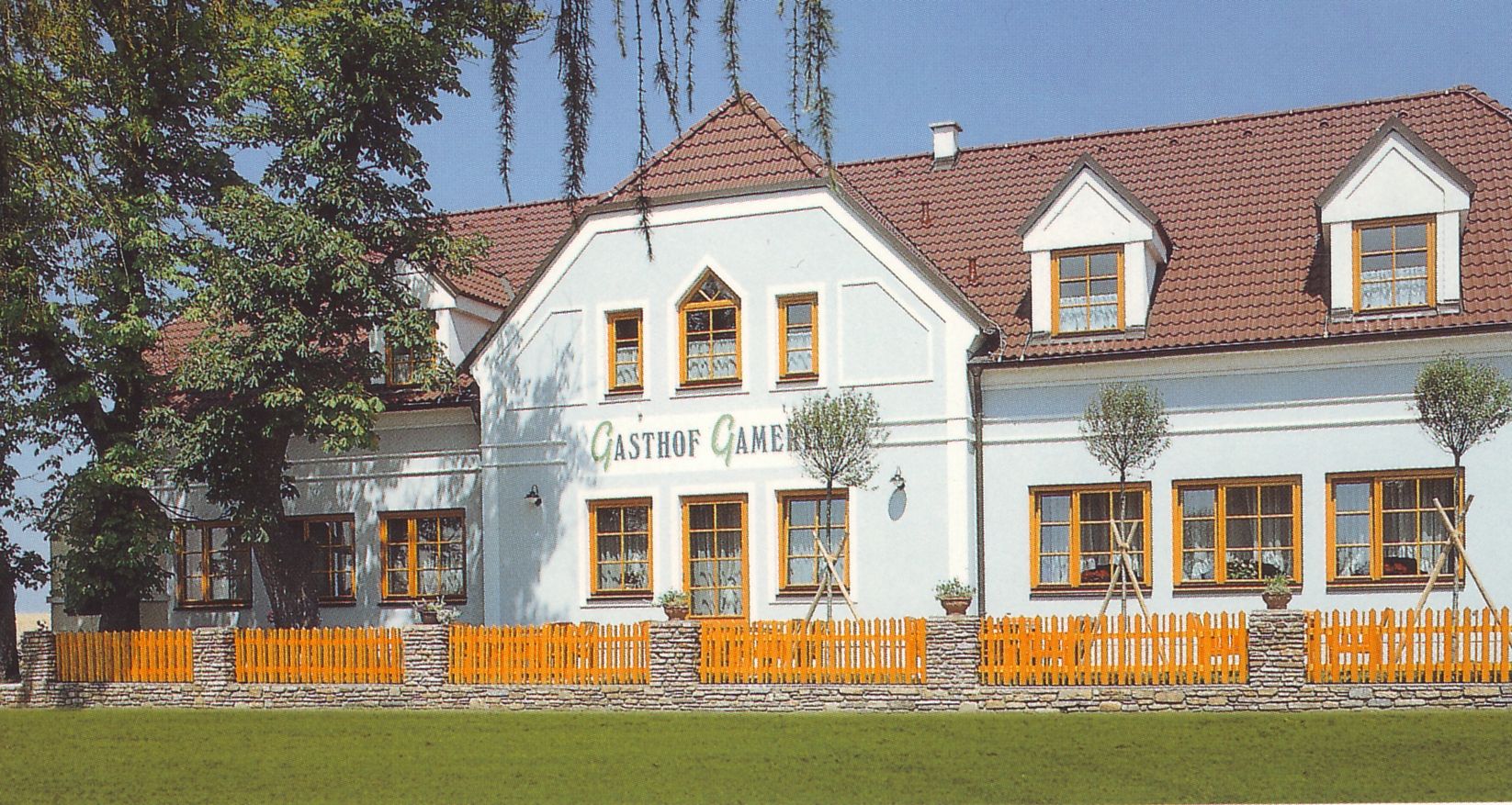 Tradičný hostinec s bielou fasádou a červenými škridlami, obklopený stromami a dreveným plotom.