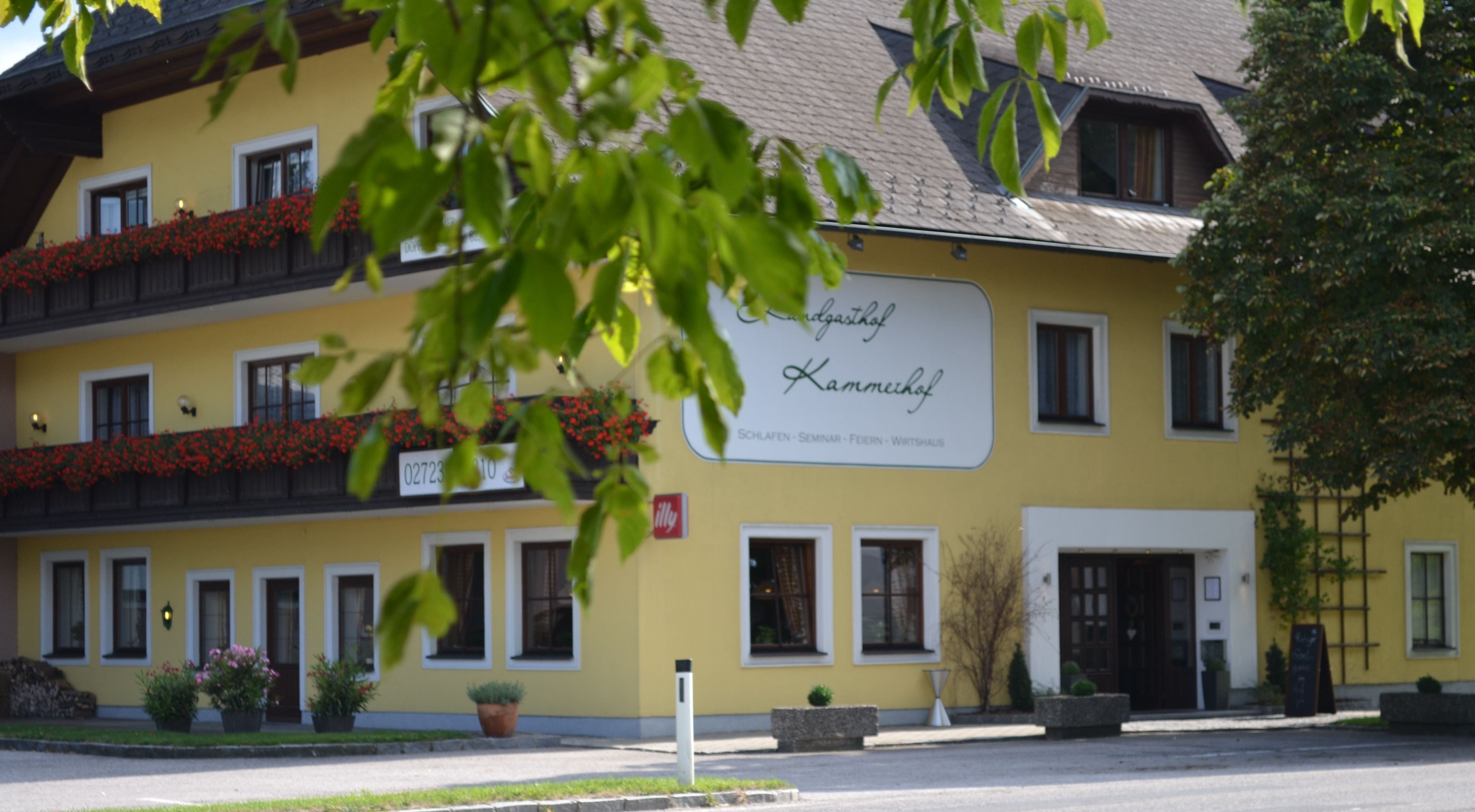 Žltá budova s nápisom "Landgasthof Kammerhof", kvetináče a stromy v popredí.