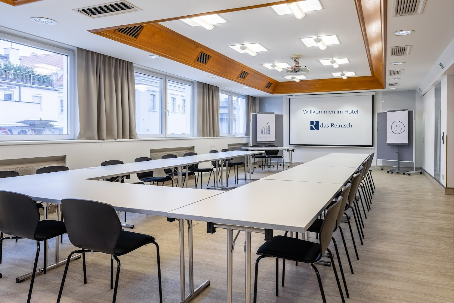 Seminárna miestnosť v hoteli Das Reinisch so stolmi, stoličkami a prezentačným plátnom.