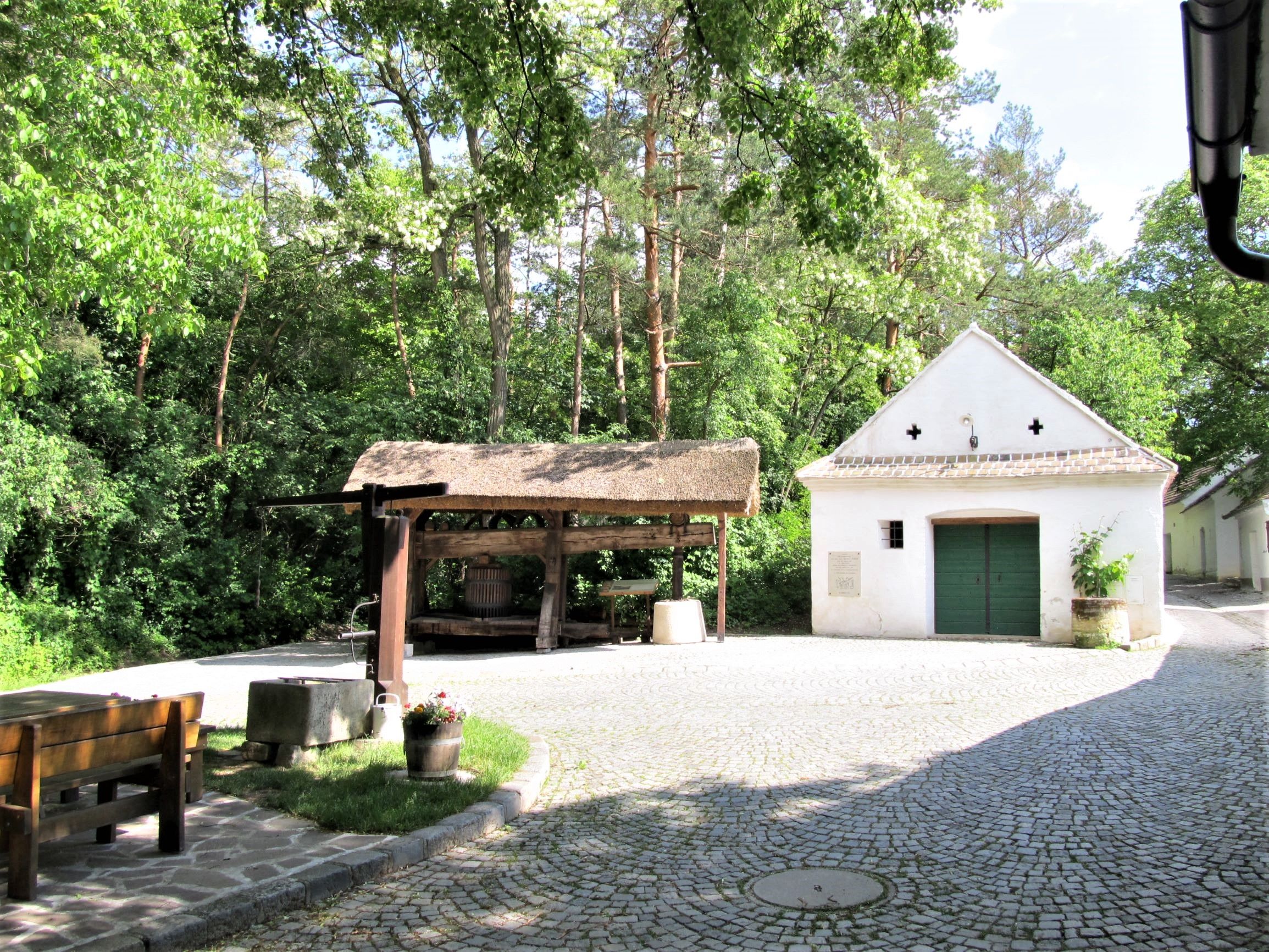 Tradičná vínna pivnica so starým lisom na víno a malou bielou budovou v pozadí, obklopená stromami.