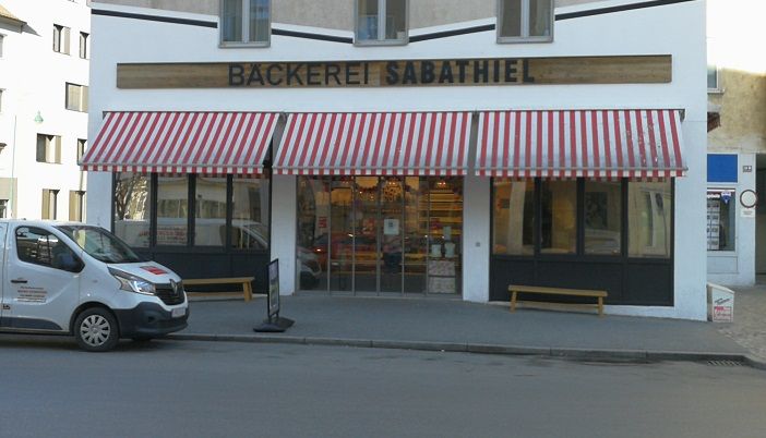 Exteriérový pohľad na pekáreň Sabathiel s červeno-bielo pruhovanou markízou a dodávkou vpredu.