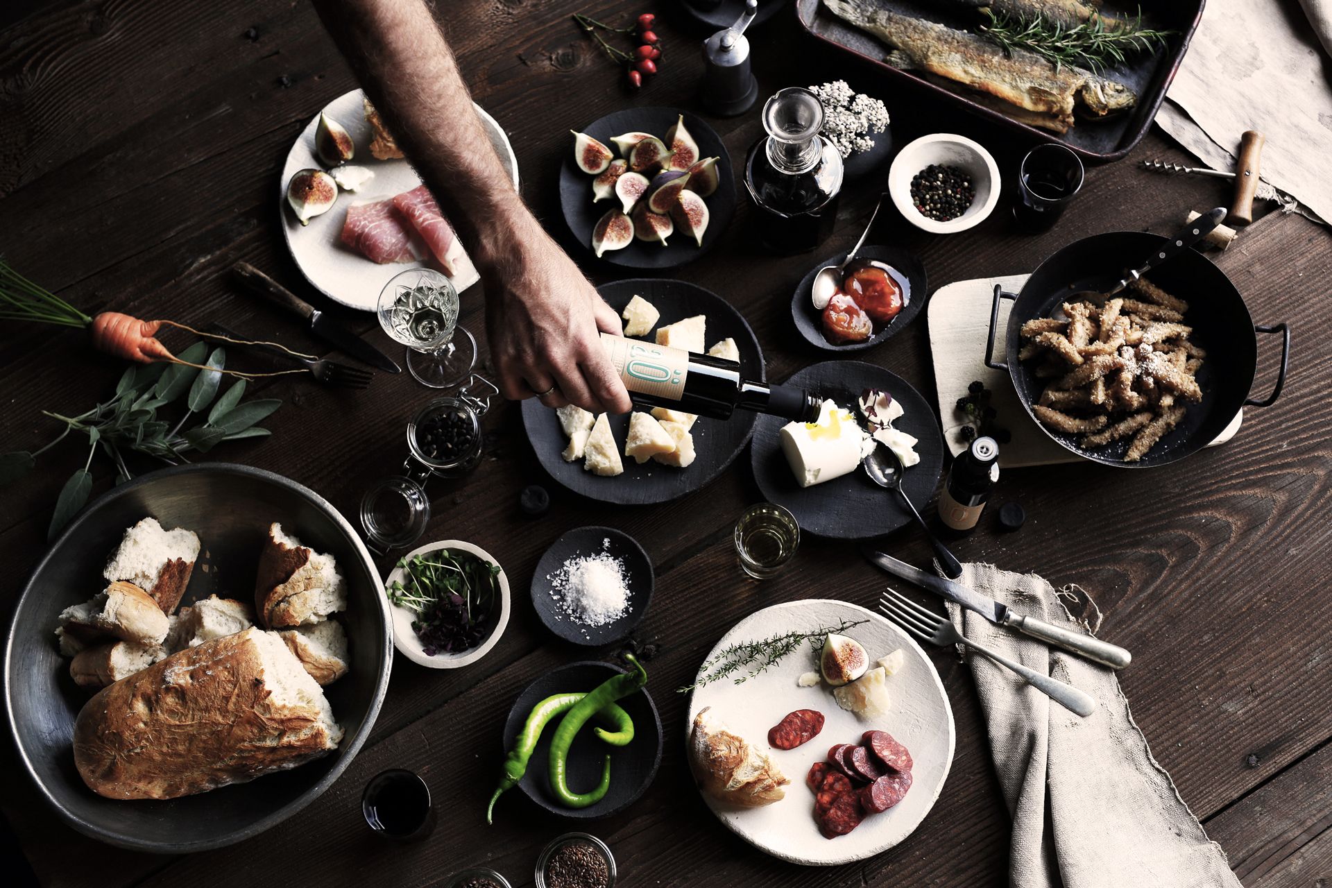 Rustikálny stôl s rôznymi jedlami vrátane chleba, syra, fíg, klobásy a rýb. Ruka leje olej na tanier.