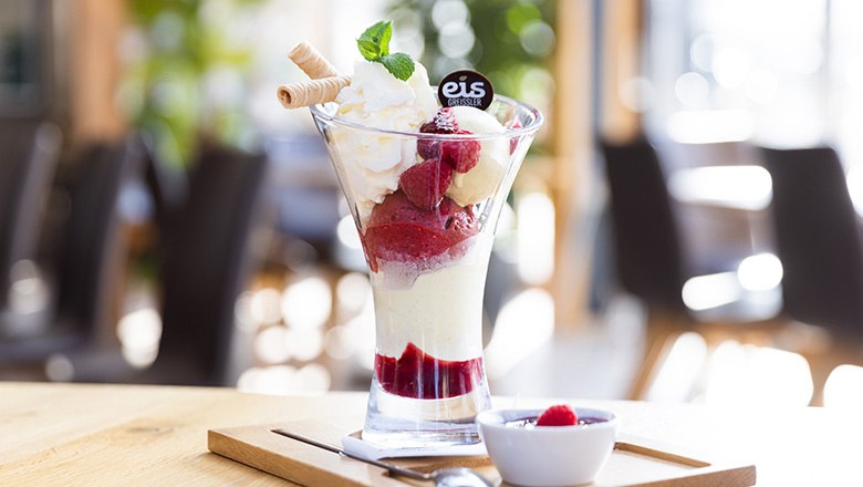A sundae with fruit ice cream, whipped cream, waffles and raspberries on a wooden board., © Blochberger Eisproduktion GmbH