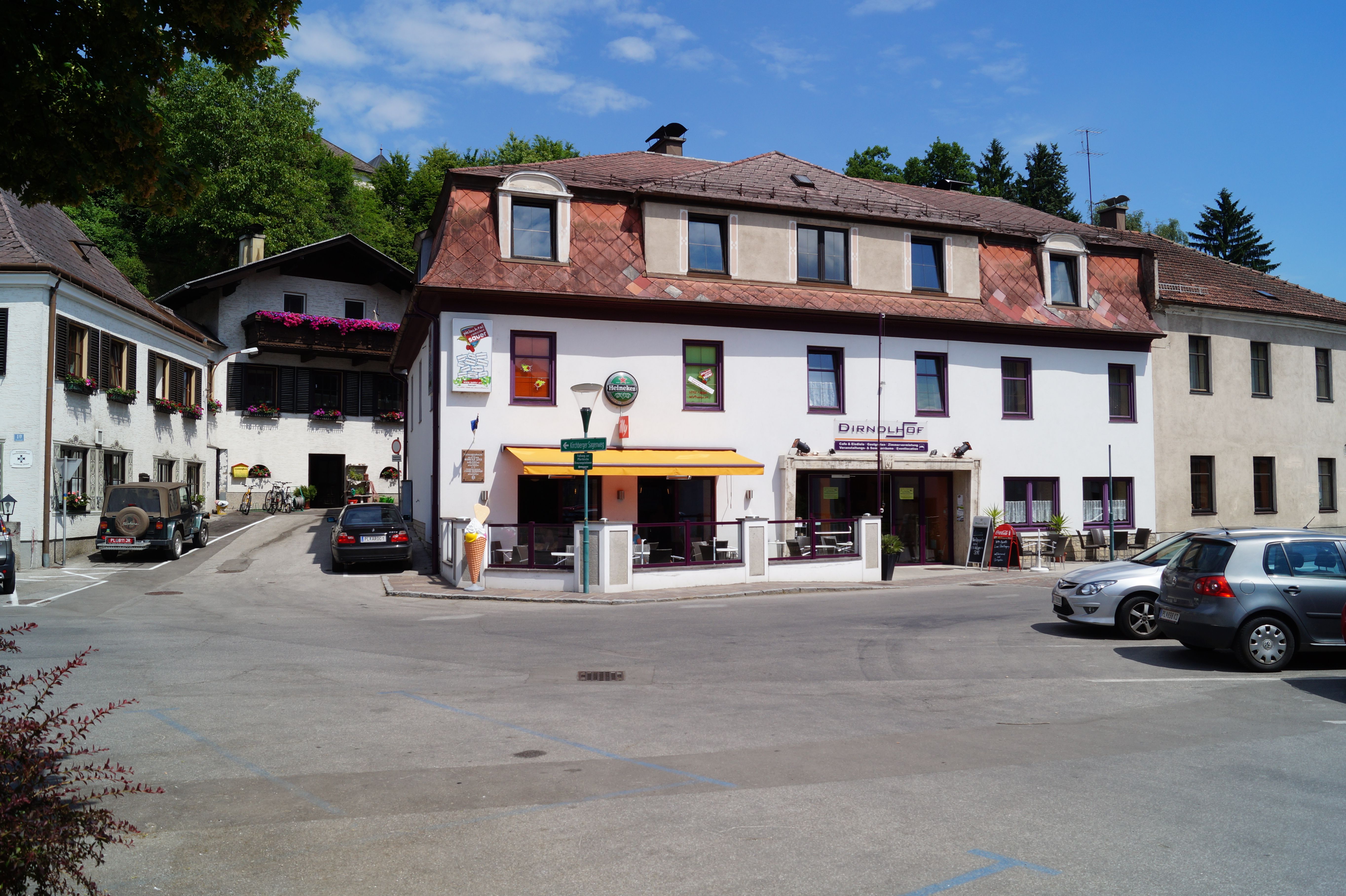 Budova s označením "Dirndlhof" a terasa so slnečníkom. Autá sa parkujú na parkovisku pred ňou.
