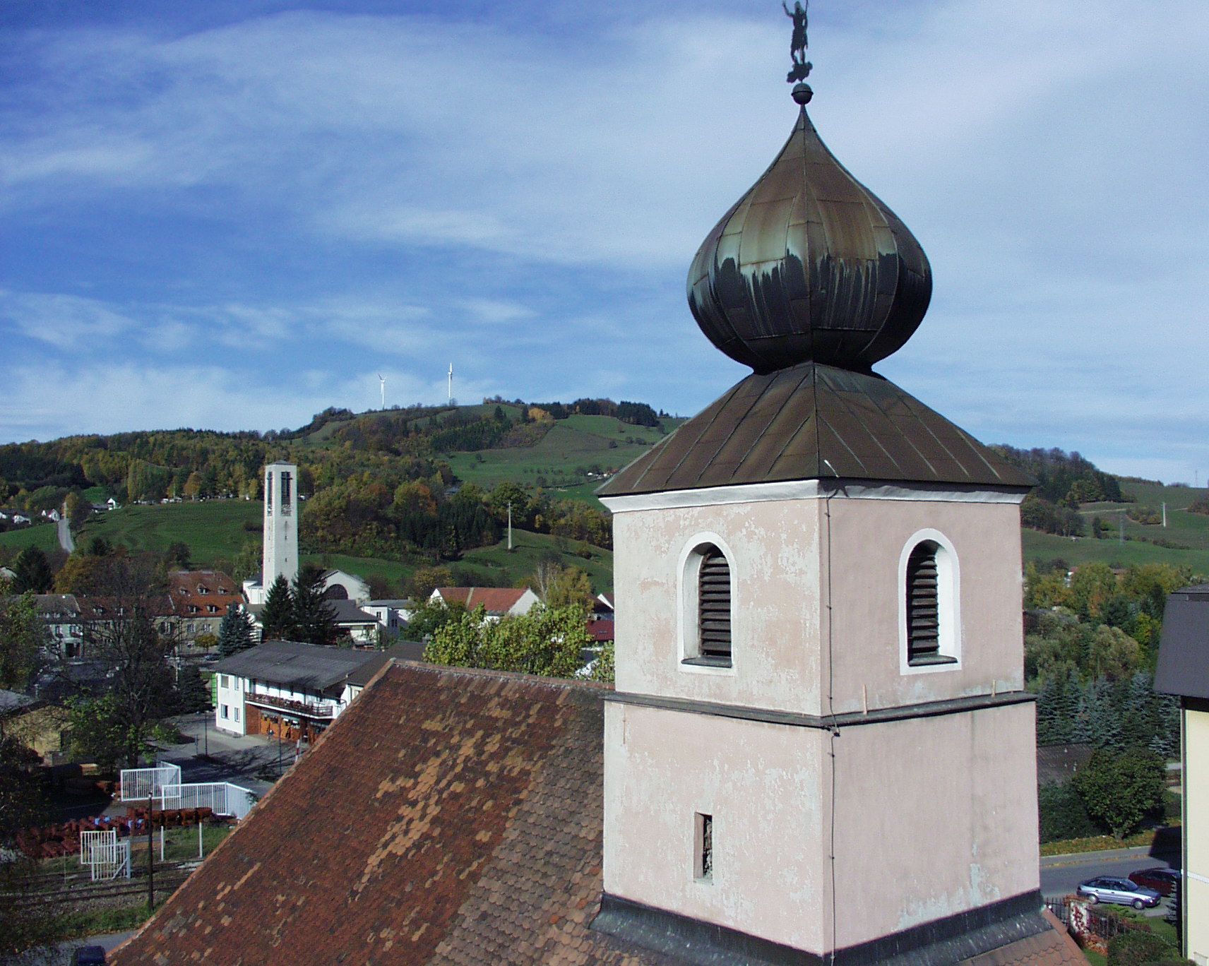 Kostolná veža s cibuľovou strechou pred kopcovitou krajinou a veternými turbínami.