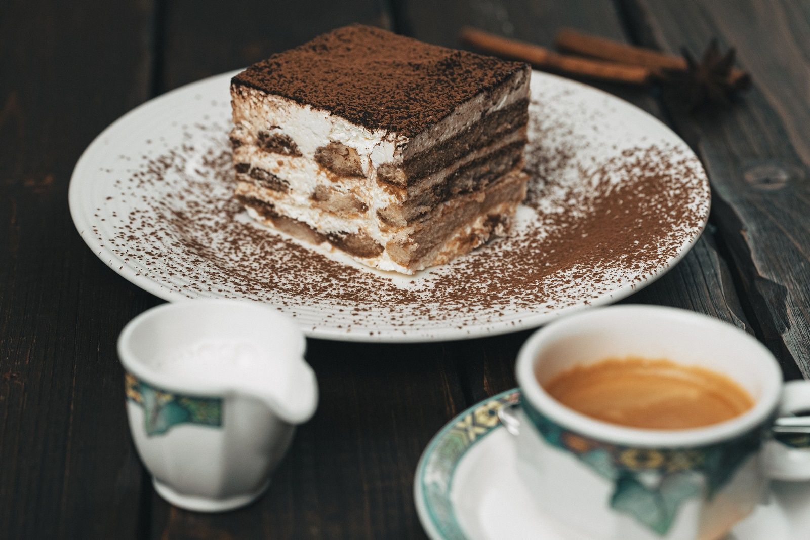 Na drevenom stole tanier s kúskom tiramisu, šálka kávy a vedľa nej kanvička s mliekom.