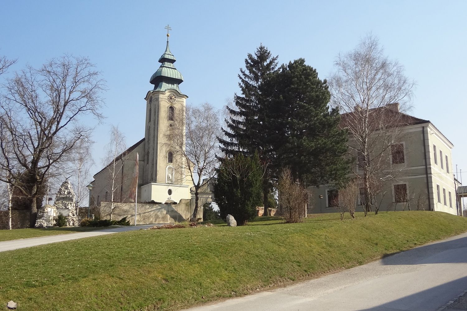 Kostol s vežou a susednou budovou v Gnadendorfe, obklopený stromami a trávnikom.