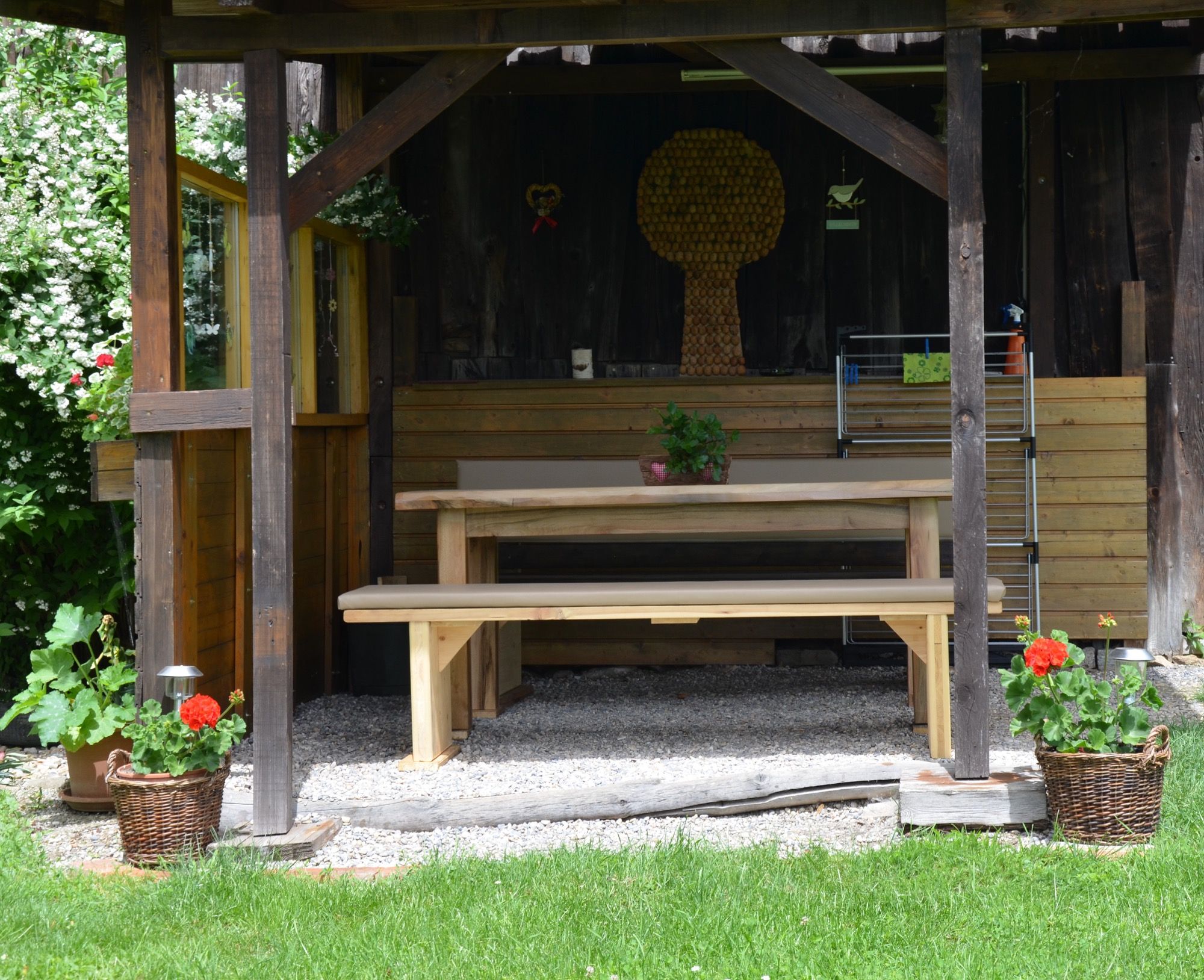 Rustikálny altánok s dreveným stolom a lavicami, obklopený rastlinami a kvetmi v košíkoch.