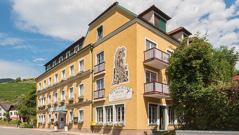 Žltá budova hotela s balkónmi a rastlinami, modrá obloha v pozadí.