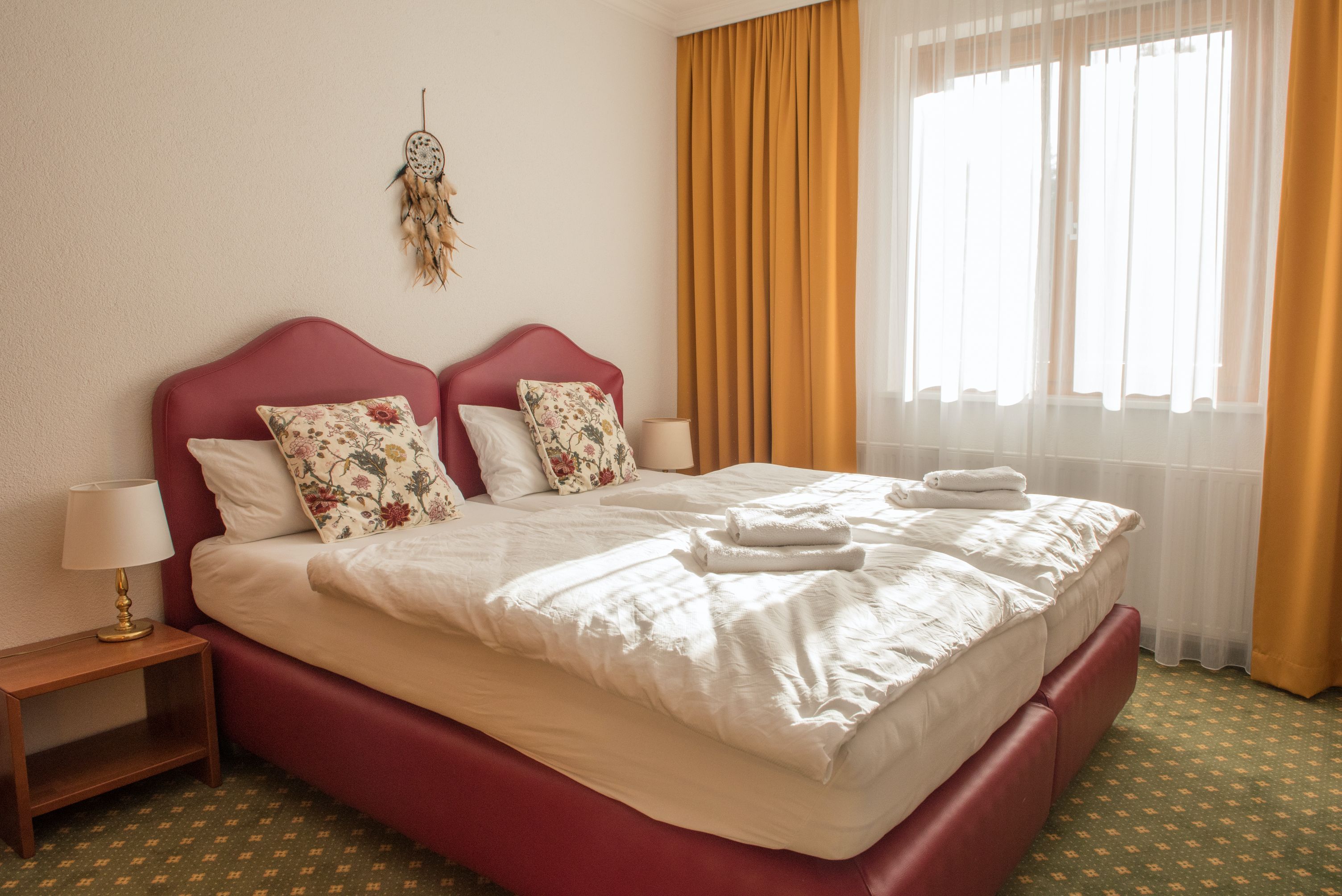 Útulná dvojlôžková izba s dvoma posteľami, žltými závesmi a lapačom snov na stene.