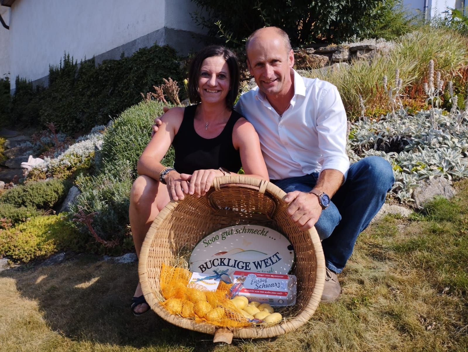 Žena a muž sedia s úsmevom v záhrade a držia košík so zemiakmi a vajíčkami.