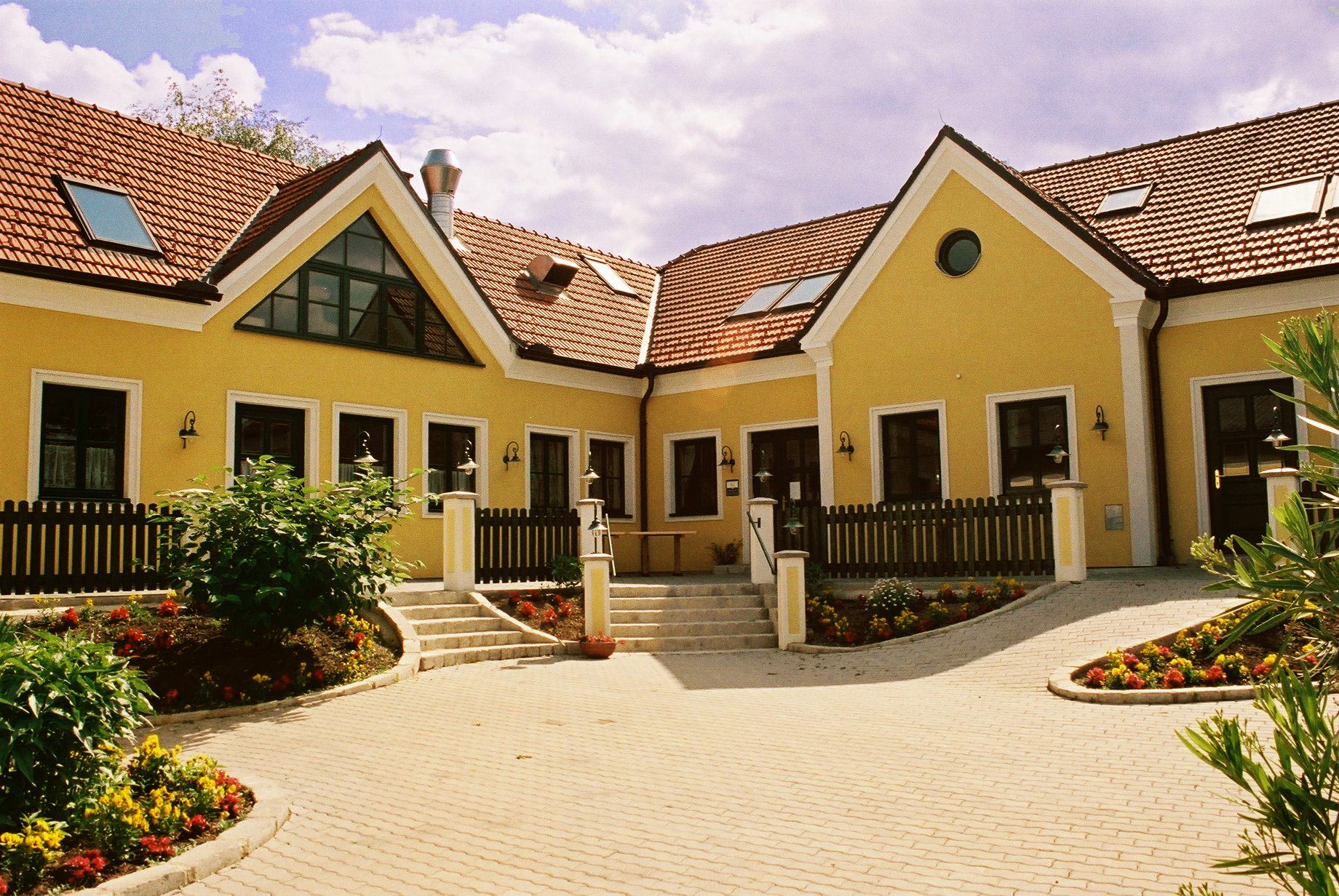 Žltá budova s červenými škridlovými strechami a záhradou.