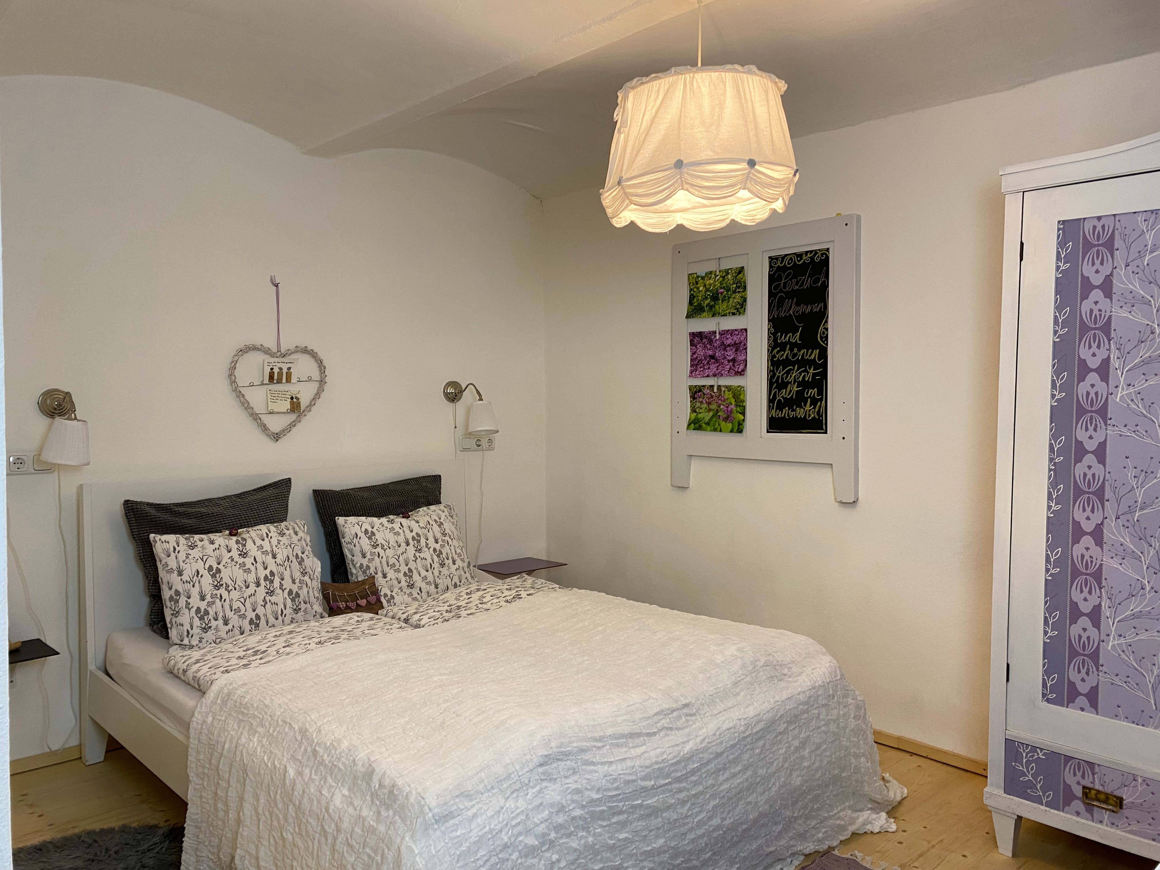 Útulná dvojlôžková izba s bielou posteľou, ozdobnou poličkou so srdcom a fialovou skriňou.