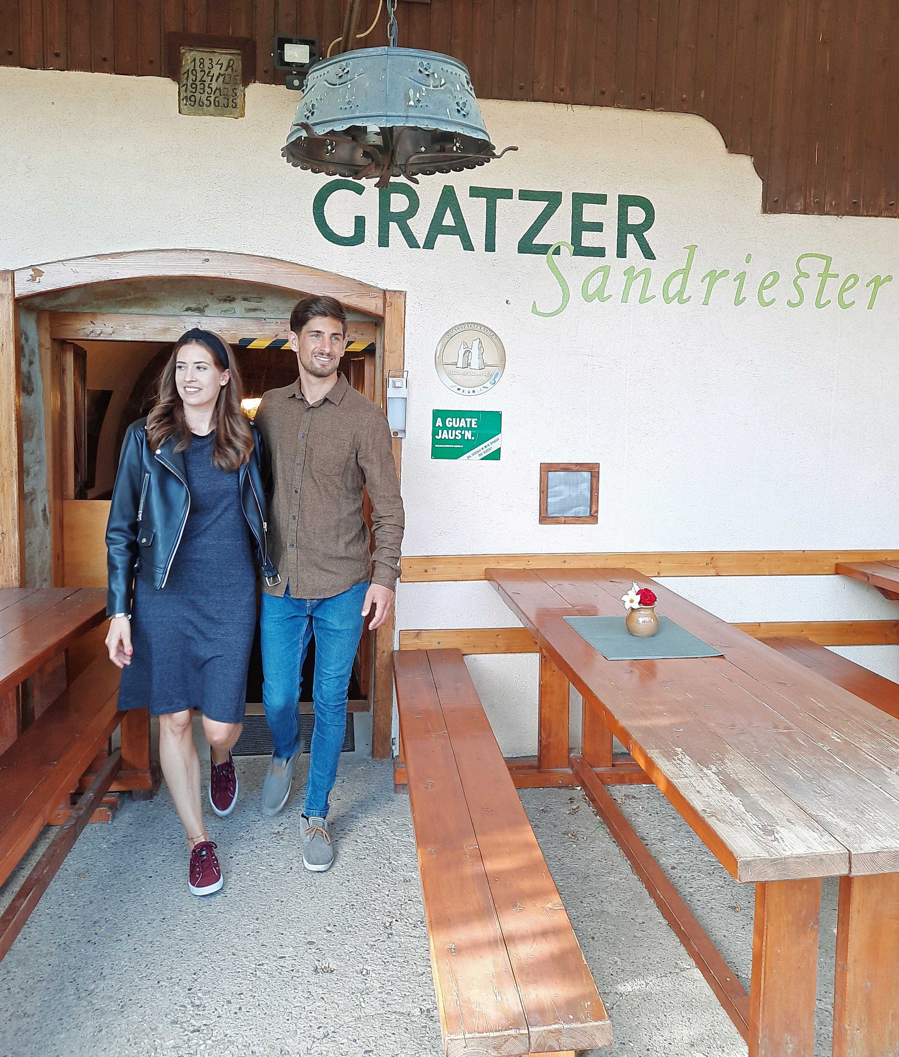 Muž a žena odchádzajú z vínneho šenku Gratzer-Sandriester. Usmievajú sa a prechádzajú okolo drevených lavičiek.