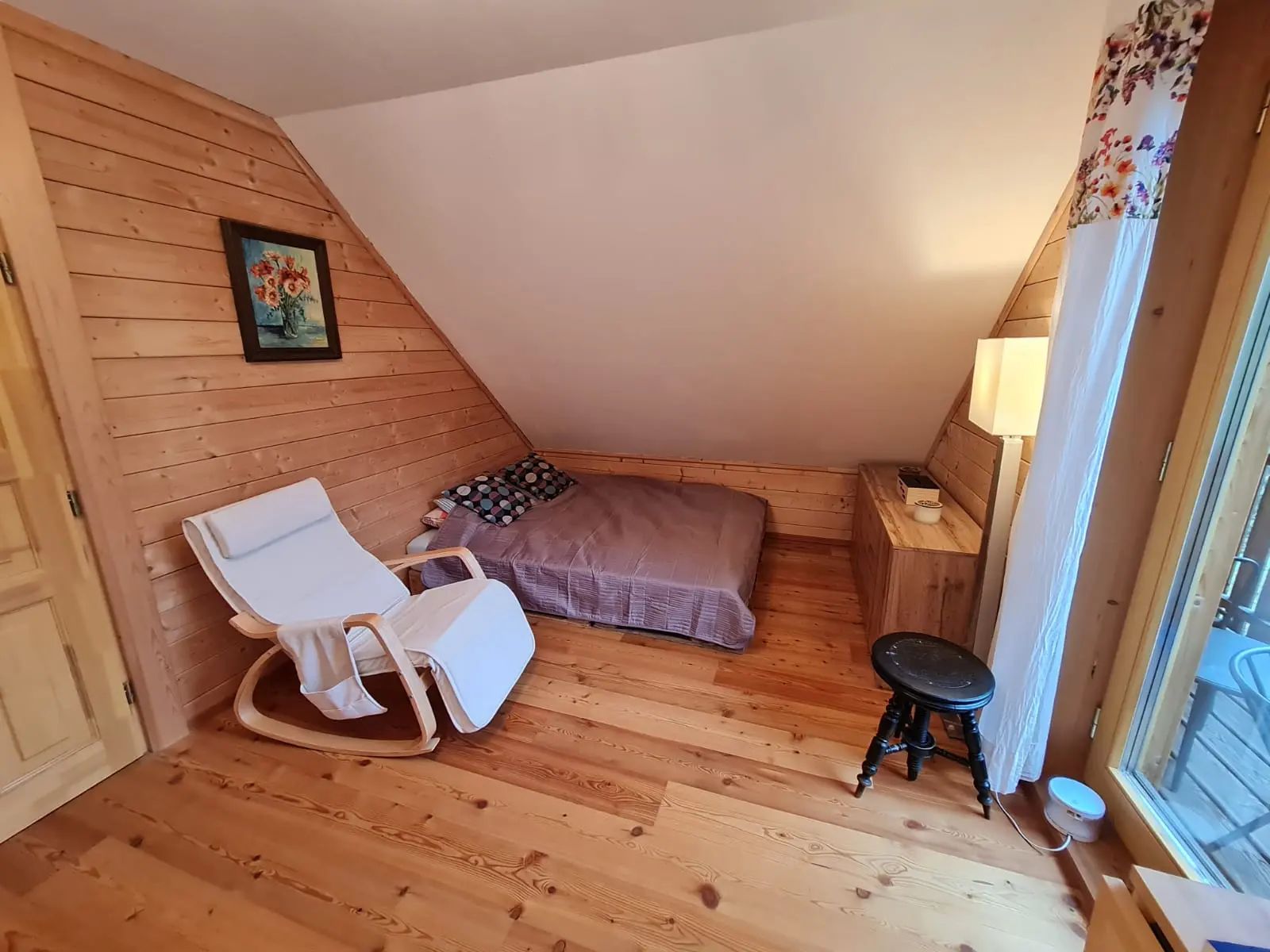 Útulná izba s drevenými stenami, posteľou, bielym hojdacím kreslom a malým stolíkom.