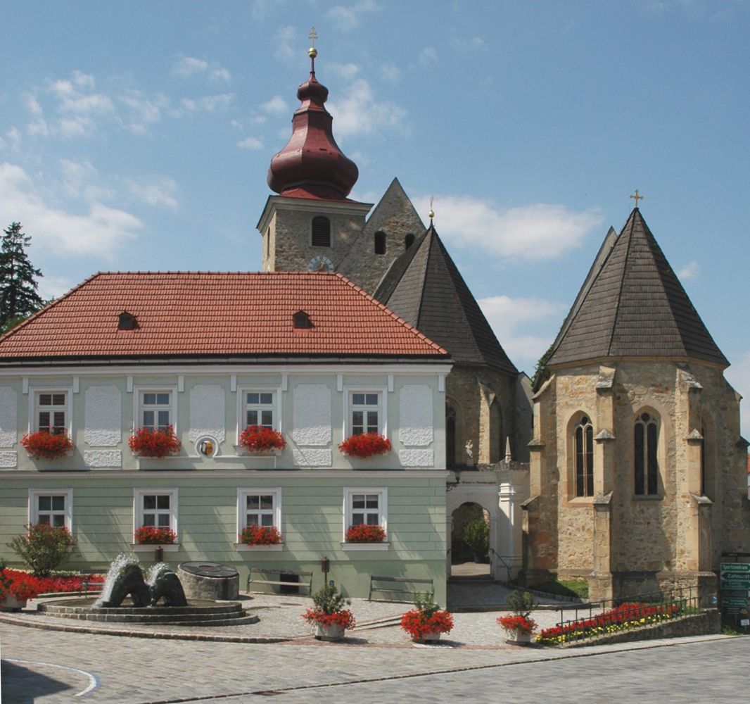 Historická budova s červenou strechou a kostolom v pozadí na námestí Maria-Anzbach.