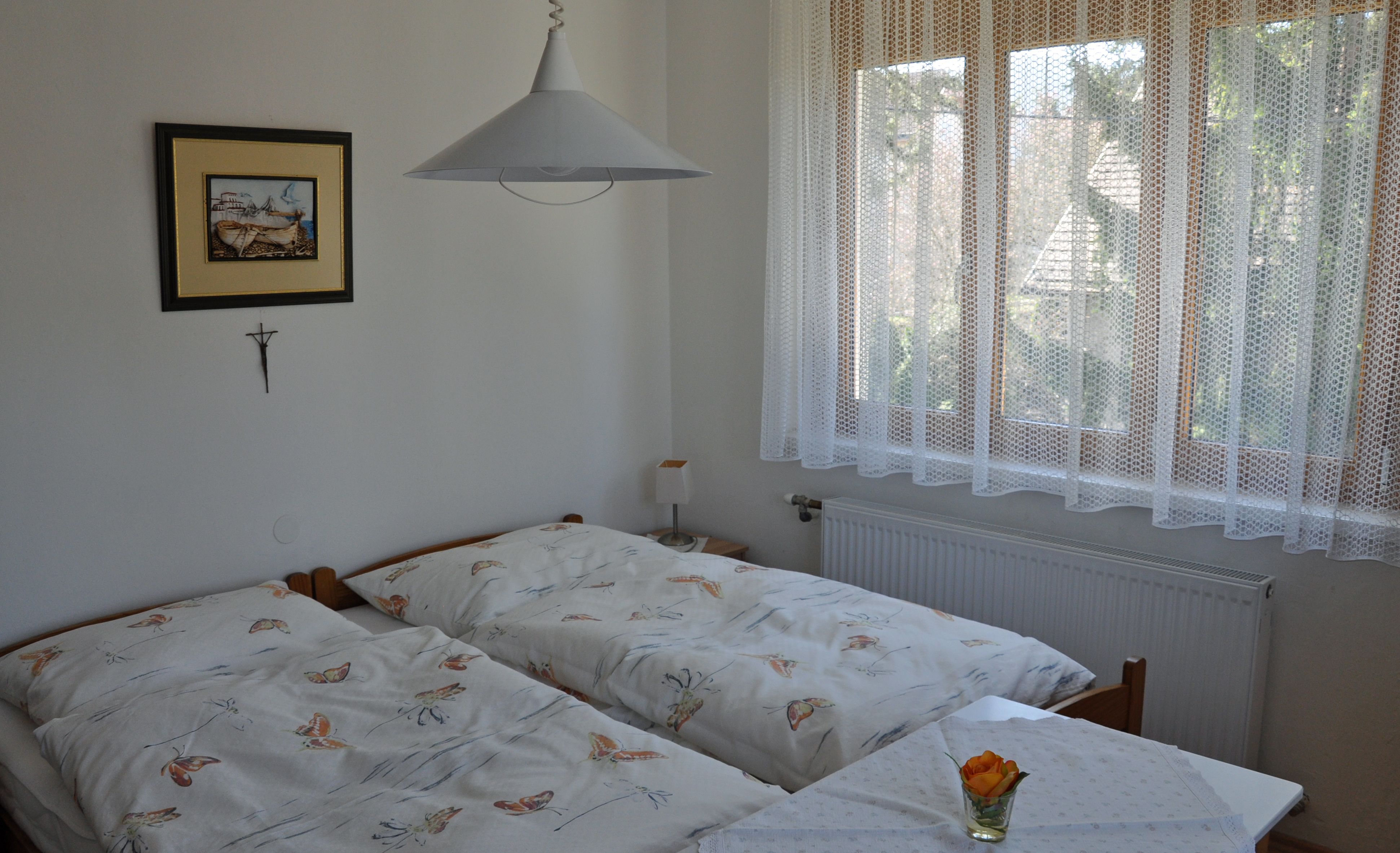 Útulná spálňa s dvoma samostatnými posteľami, obrazom na stene a oknom s čipkovaným závesom.