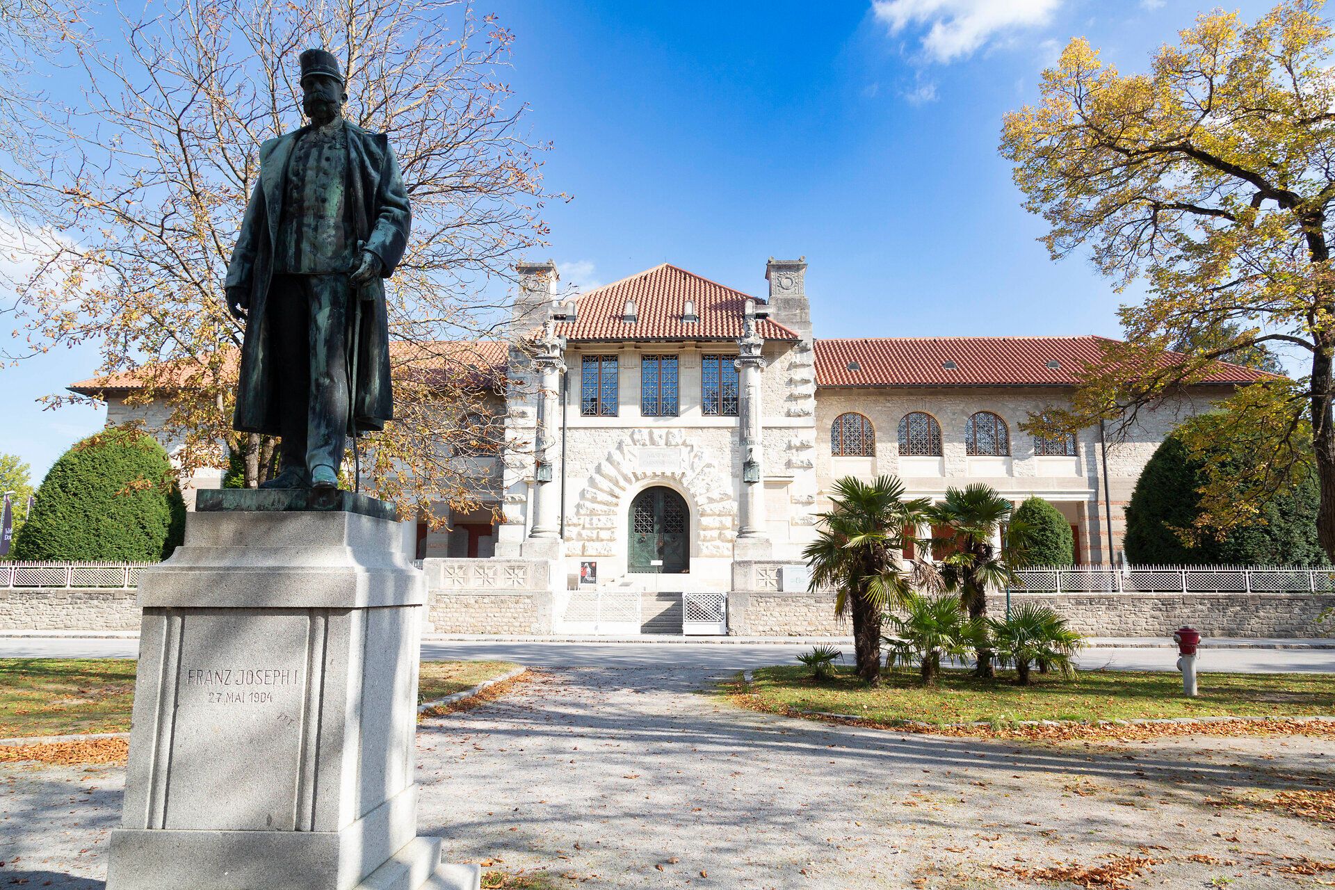Majestátna socha hrdo stojí pred pôsobivou budovou, obklopená farebným jesenným lístím. Jemné slnečné lúče kúpu scenériu v hrejivom svetle a pozývajú vás objavovať krásy okolia.