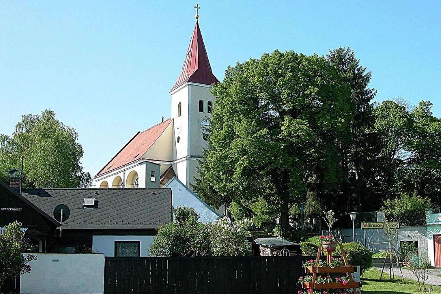 Kostol s červenou strechou a vežou, obklopený stromami a domami.