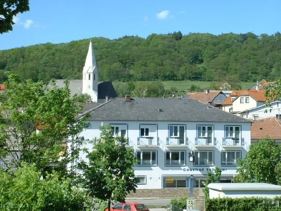 Dvojposchodová budova s nápisom "Gasthof Schauer" pred zalesneným kopcom a kostolom so špicatou vežou.