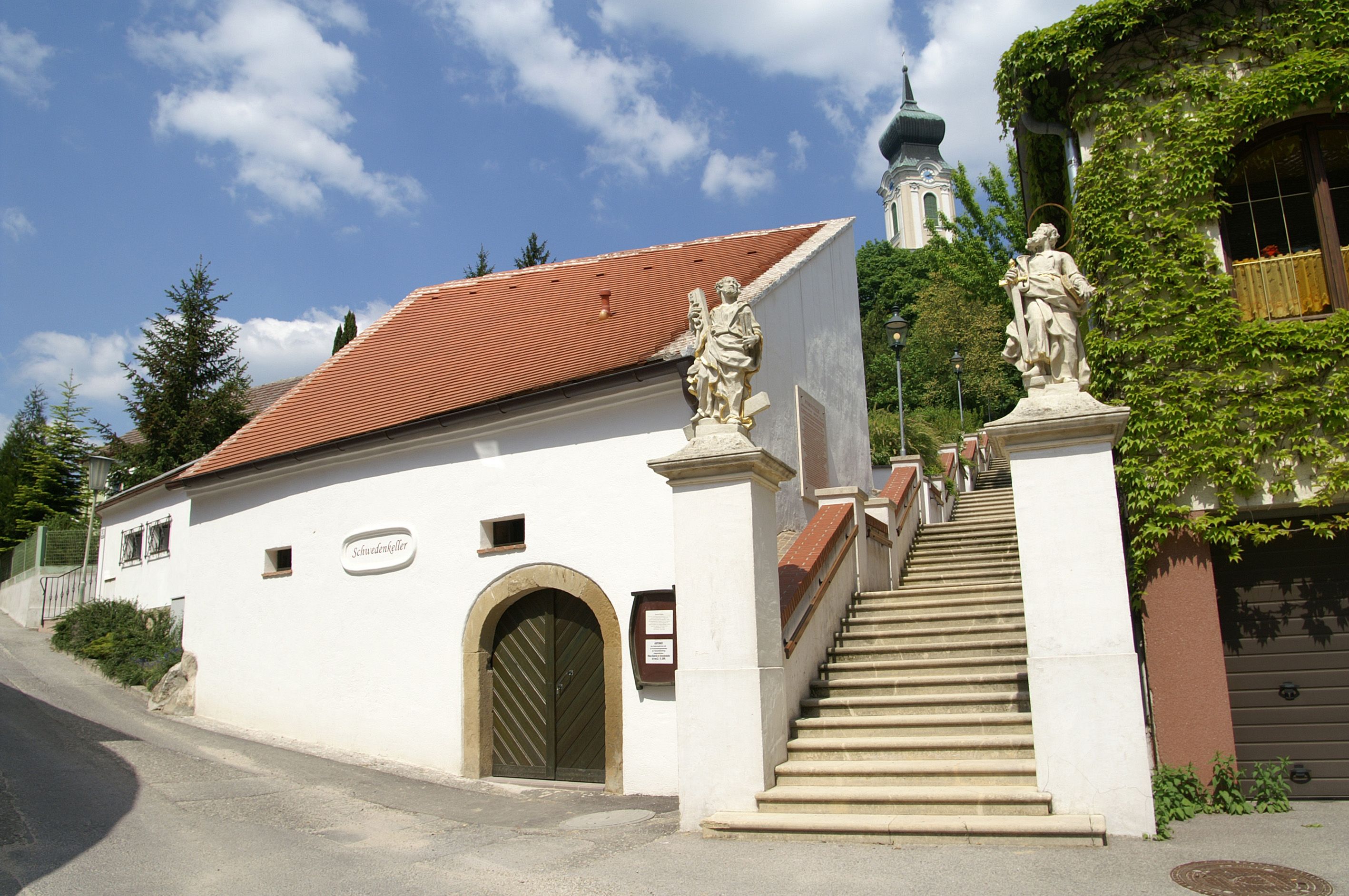 Biela budova s červenou strechou a schodiskom so sochami, v pozadí kostol.
