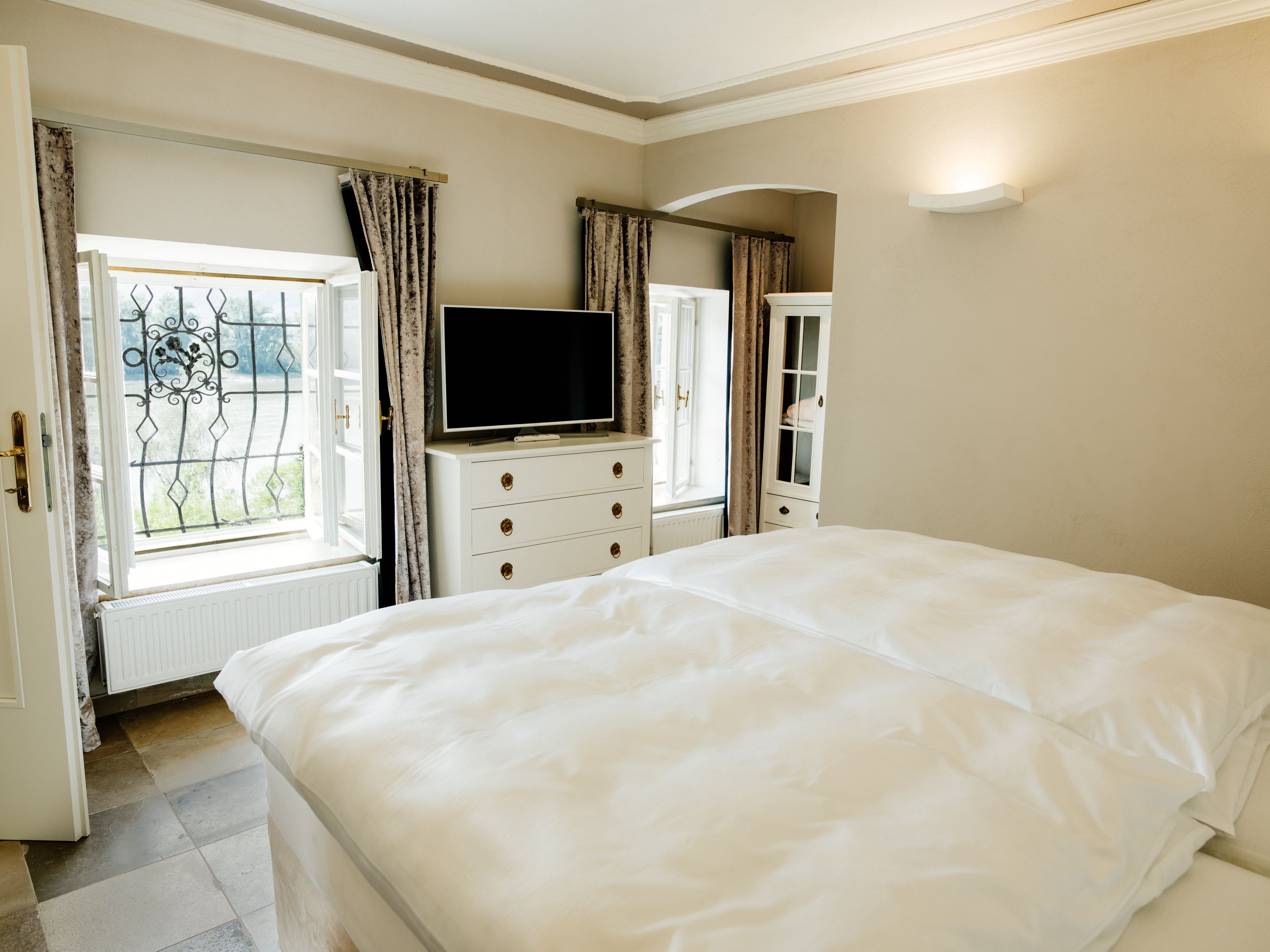 Svetlá spálňa s manželskou posteľou, TV, komodou a dvoma oknami so závesmi.