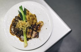 Tanier s hov&auml;dzou pečienkou, zemiakov&yacute;m gratinovan&iacute;m, p&oacute;rom a chrumkavou cibuľkou na kovovom povrchu.