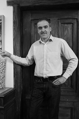 Muž v košeli stojí pred drevenými dverami a usmieva sa do kamery.