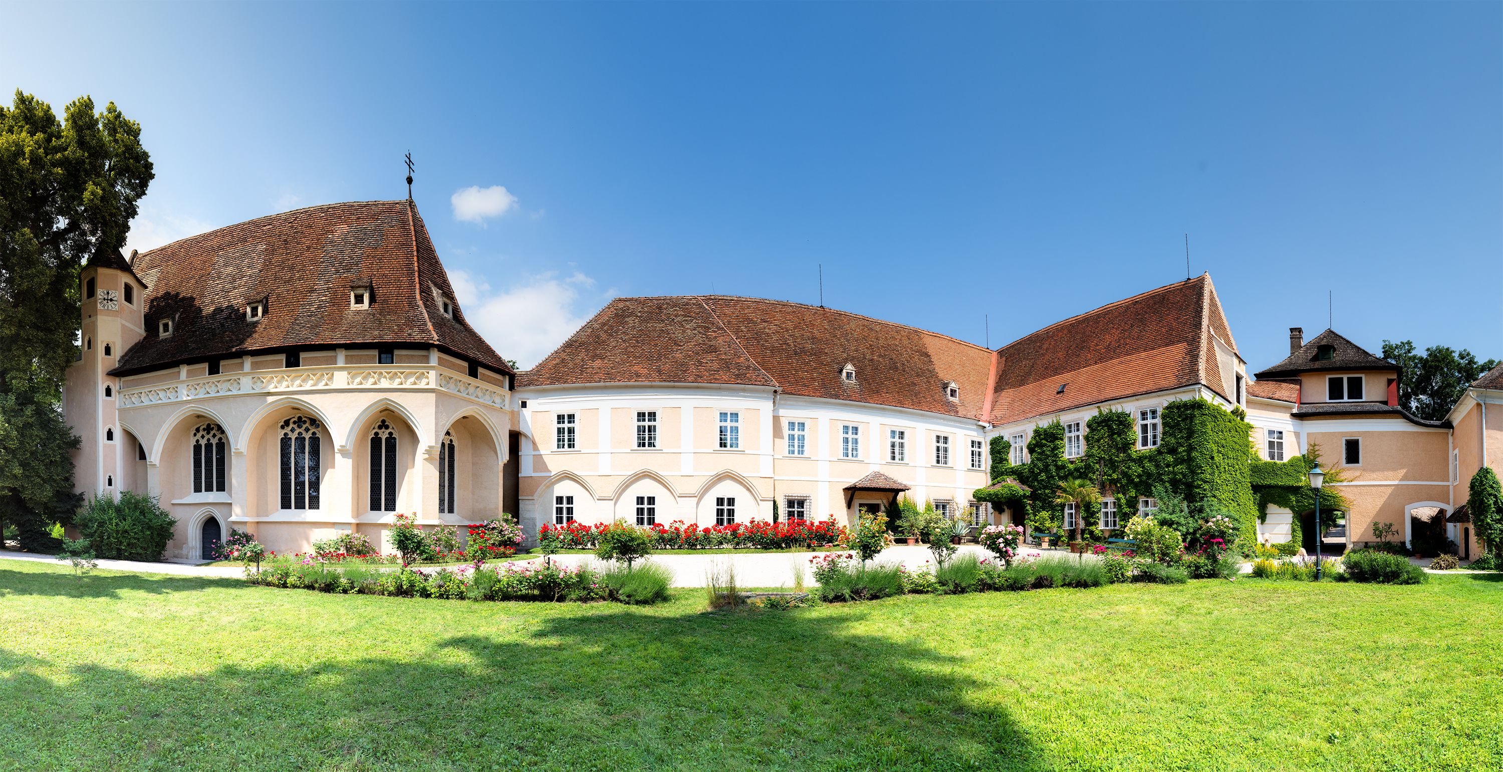 Panoramatický pohľad na zámok Schrattenthal s upravenou záhradou a kvitnúcimi ružami v popredí.