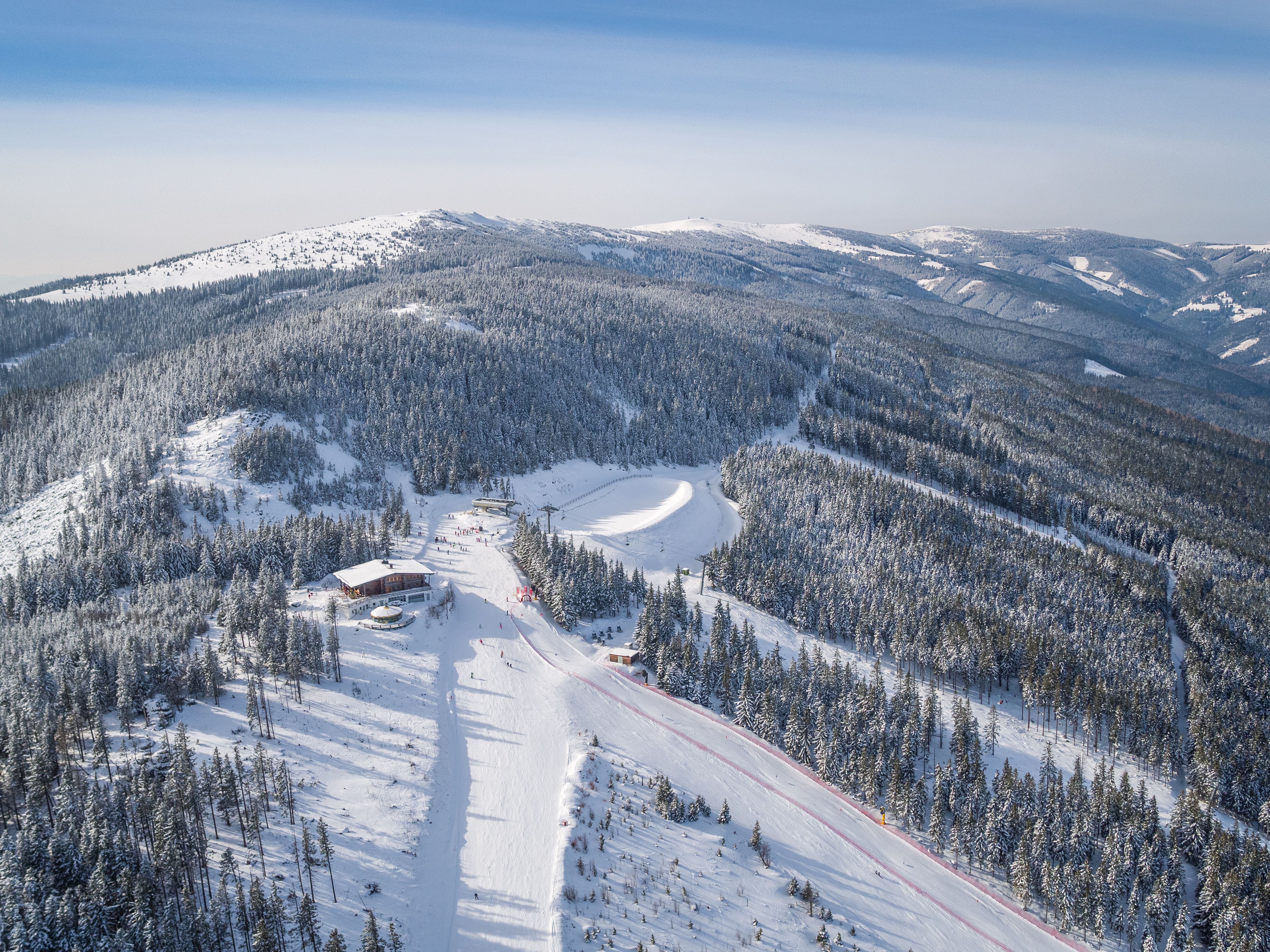 Lyžiarsky areál so sedačkovou lanovkou a lyžiarmi, obklopený zasneženými stromami a kopcami.