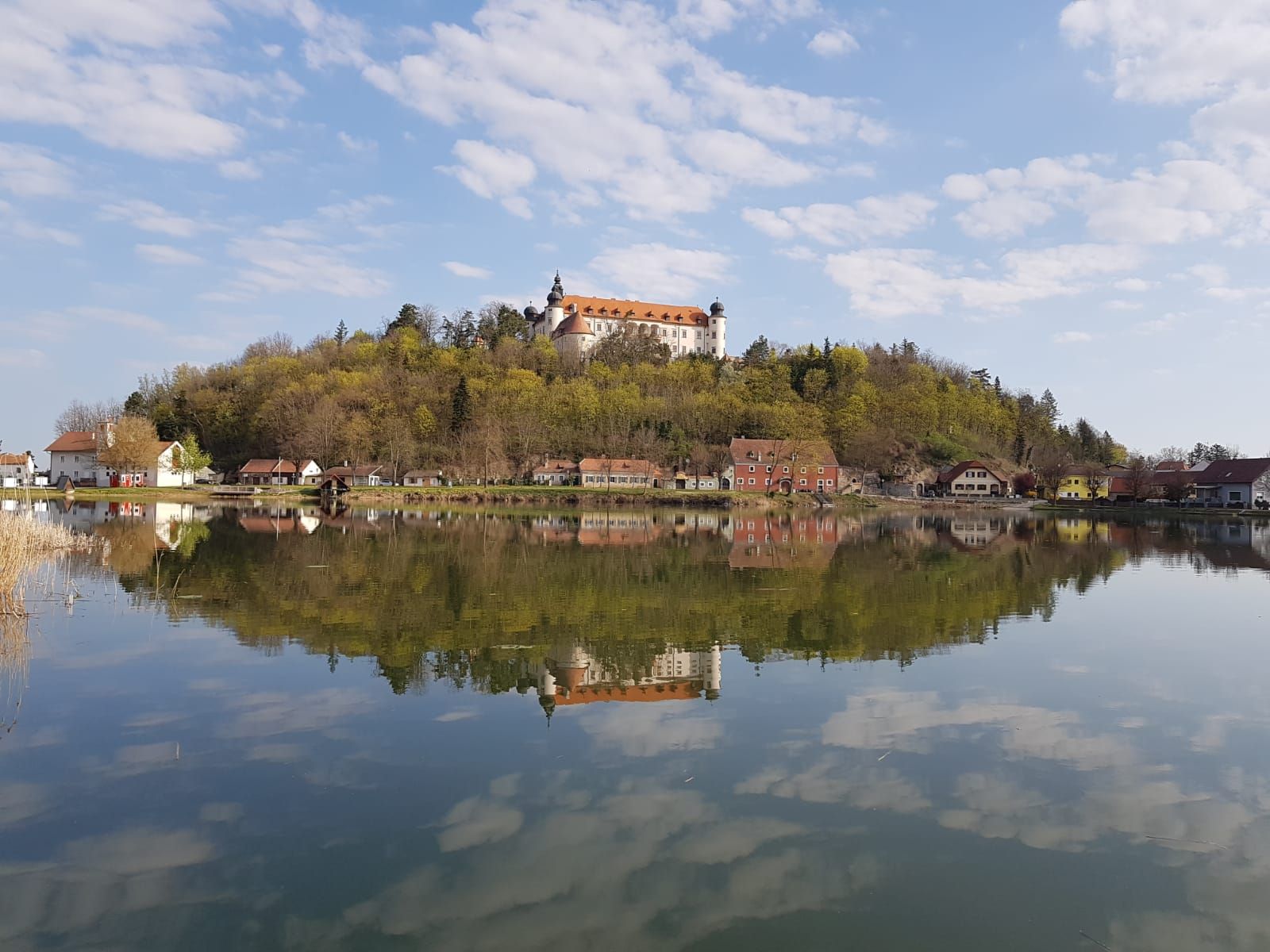 Hrad na kopci sa odráža v pokojnom jazere obklopenom stromami a malými domčekmi.