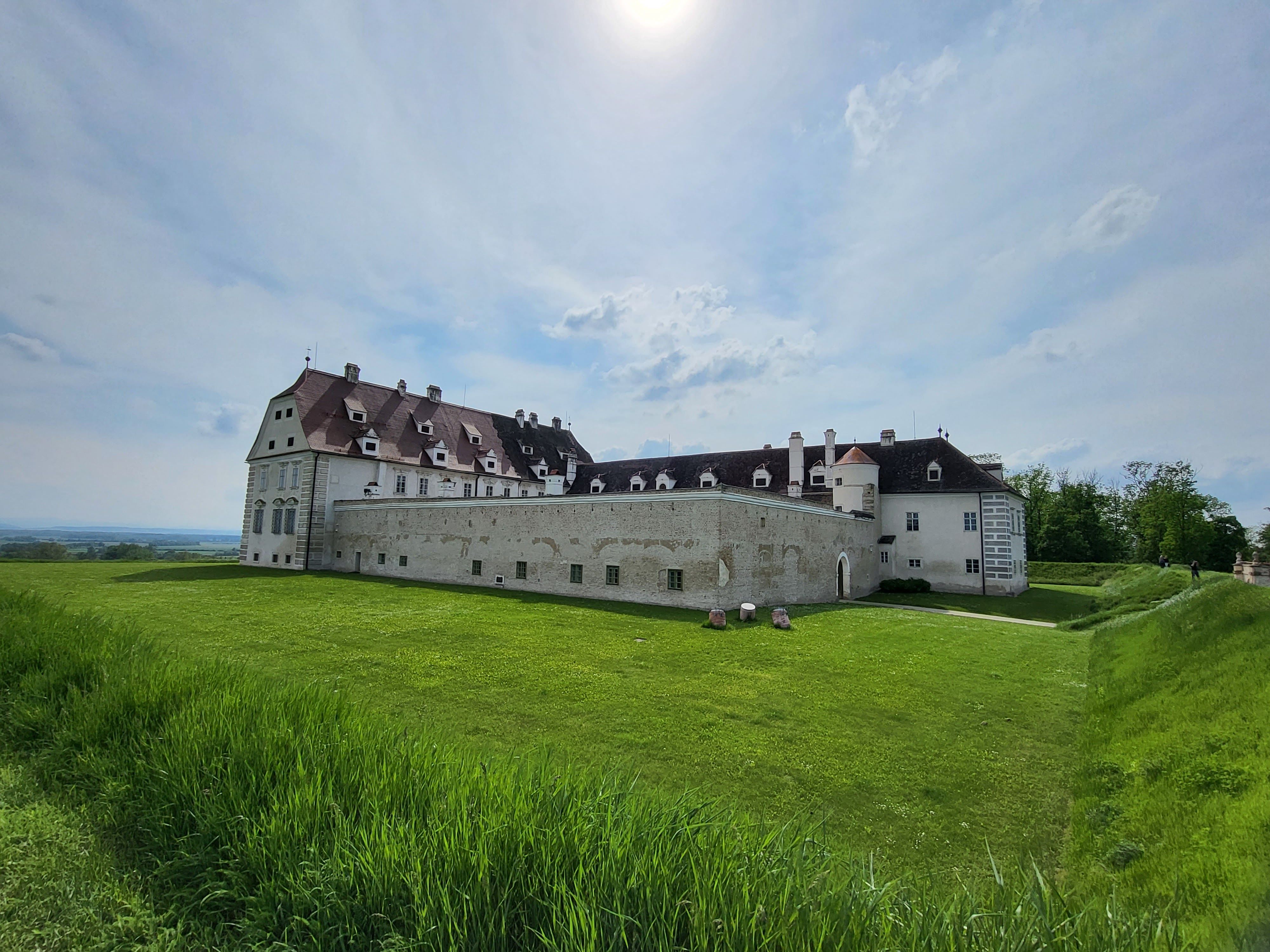Veľký historický hrad s bielymi múrmi a červenými strechami stojí na zelenej lúke pod zamračenou oblohou.