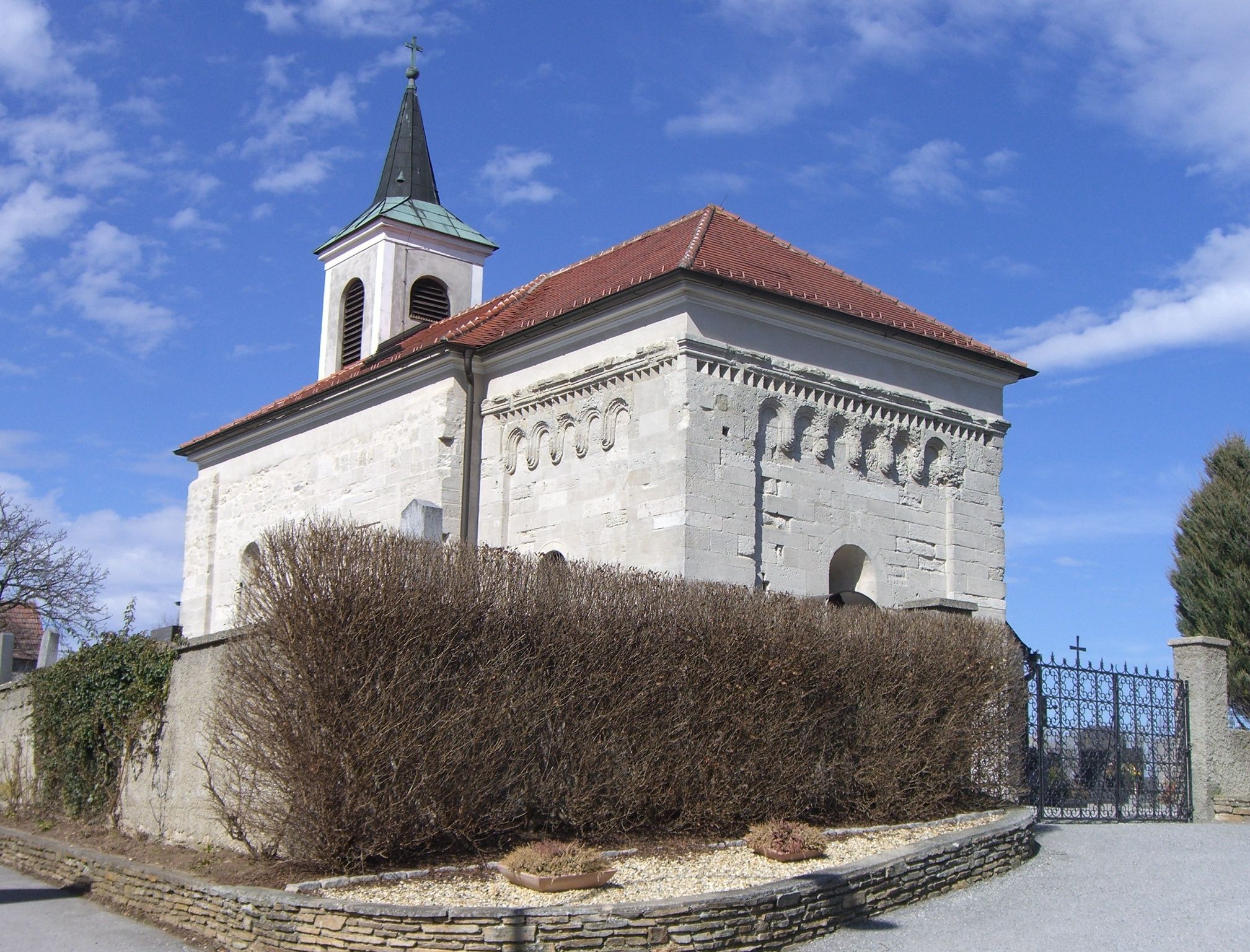 Románsky kostol so zvonicou a červenou škridlovou strechou, obklopený múrom a kríkmi.