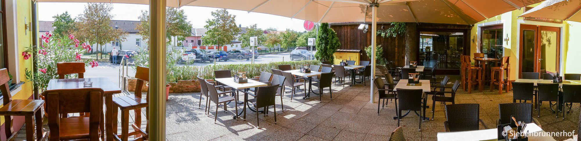 Slnečná záhrada pre hostí s prázdnymi stolmi a stoličkami, obklopená rastlinami a veľkým slnečníkom.