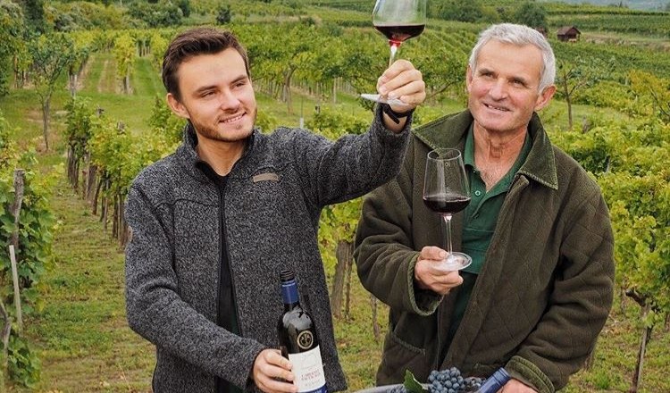 Dvaja muži vo vinici držia poh&aacute;re červen&eacute;ho v&iacute;na a usmievaj&uacute; sa. Jeden z nich drž&iacute; fľa&scaron;u v&iacute;na; v pozad&iacute; vidno vinič a kopce.