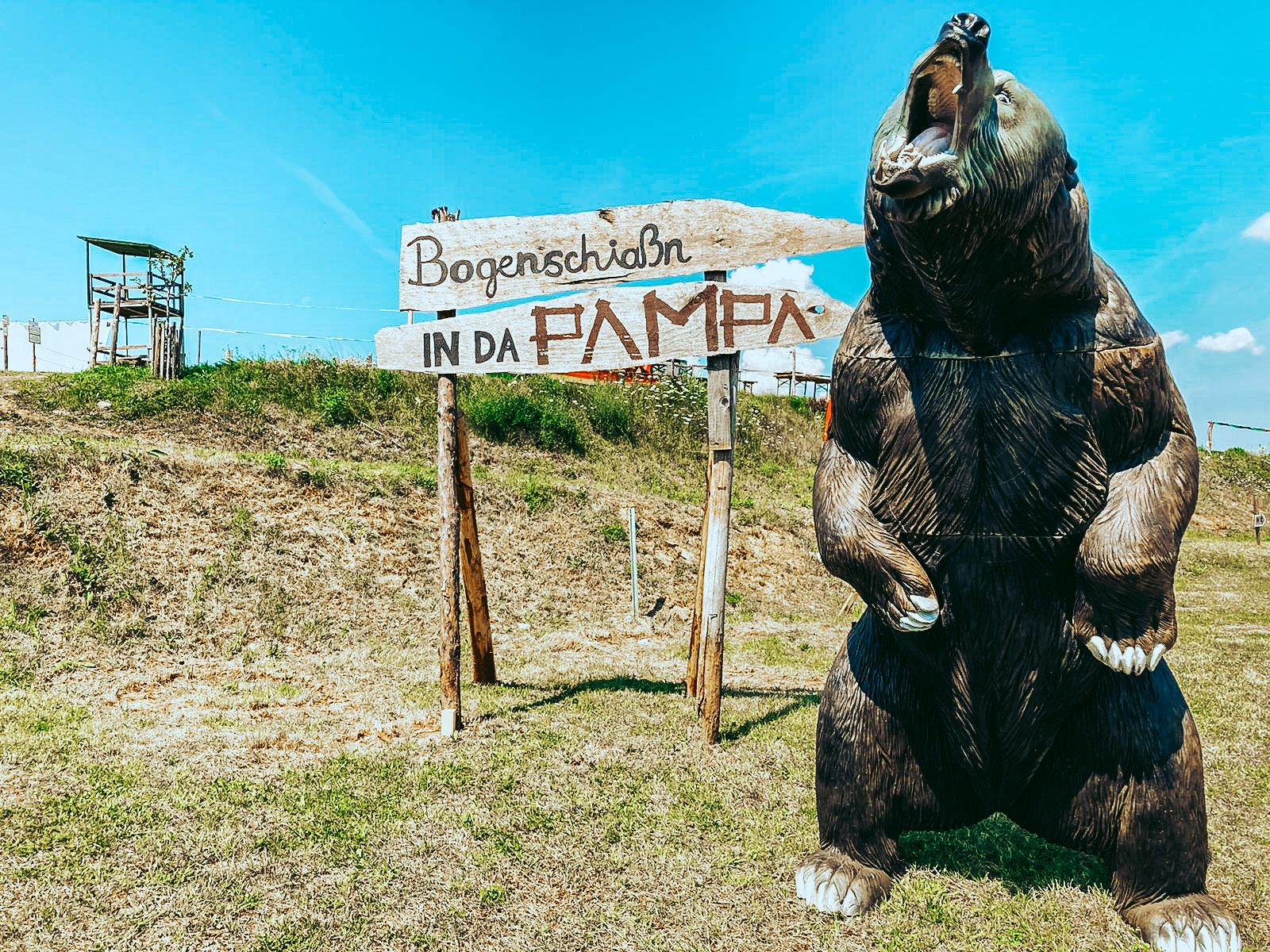 Veľký model medveďa vedľa tabule s nápisom "Bogenschiaßn in da Pampa" na lúke.
