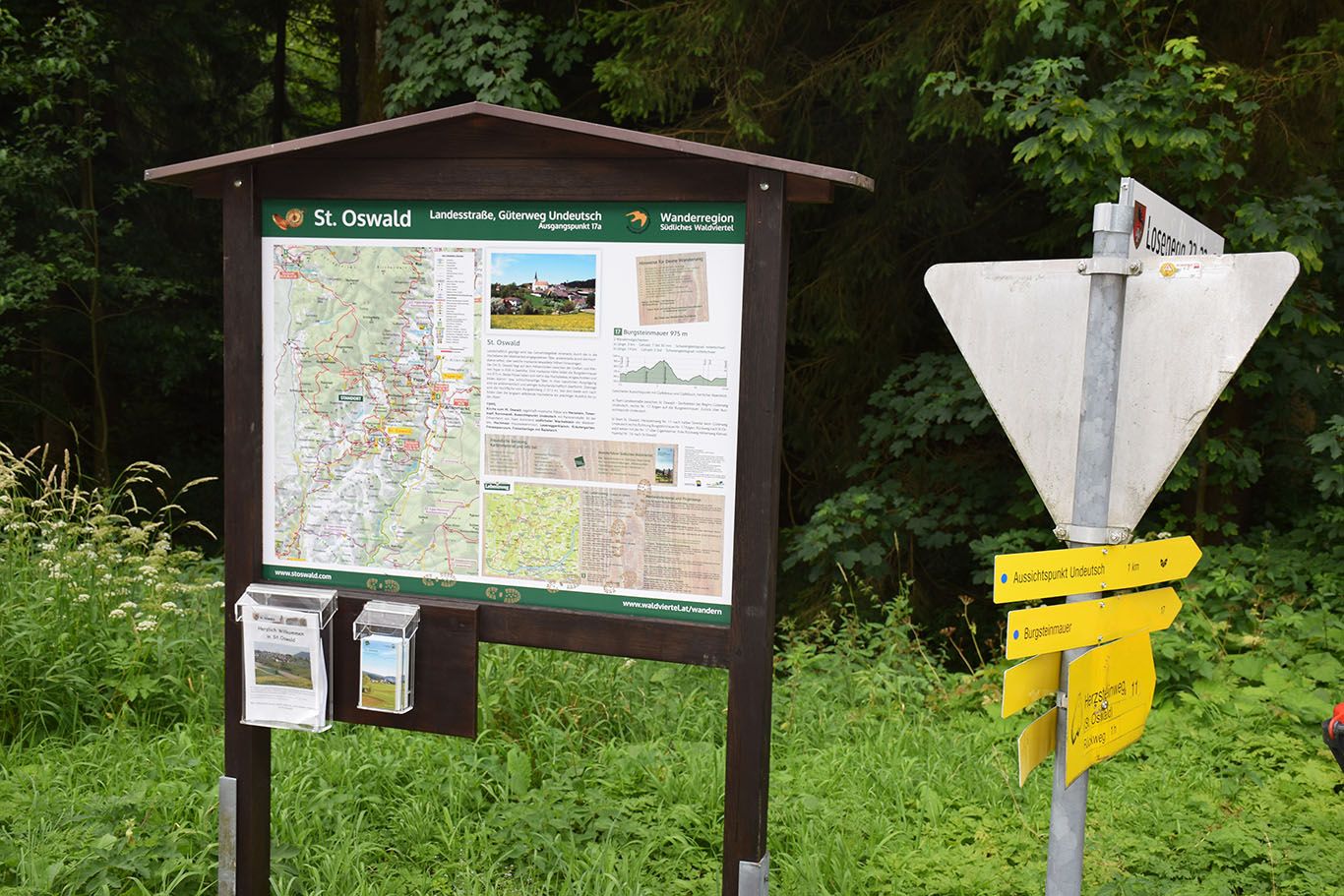 Turistický smerovník a informačná tabuľa v obci St. Oswald, obklopenej lesom a zeleňou.