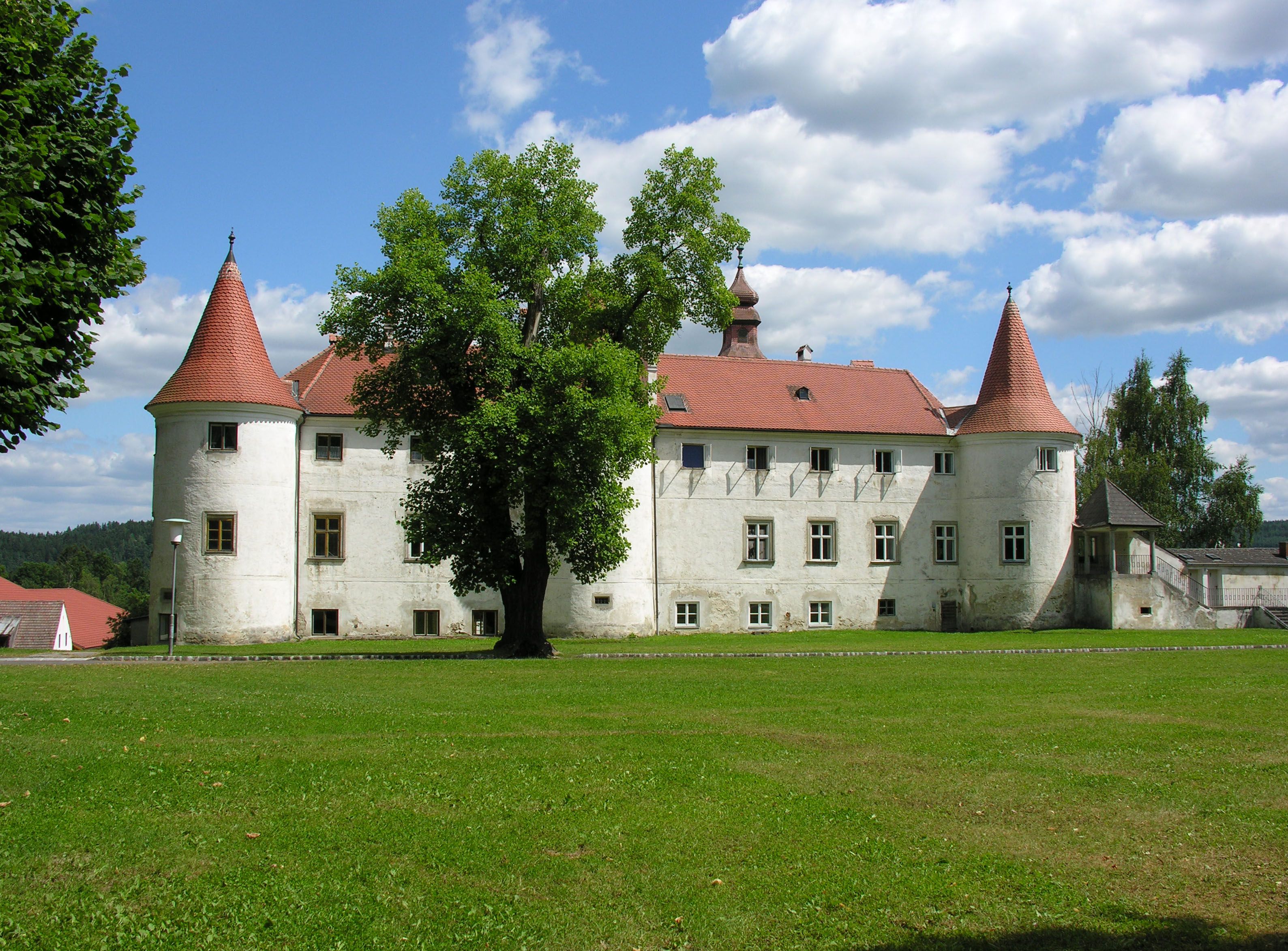 Historický hrad s červenými strechami a vežami, obklopený zelenými lúkami a stromami, pod modrou oblohou s bielymi mrakmi.