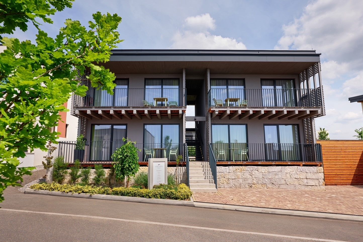 Moderný dvojpodlažný apartmánový komplex s balkónmi a schodiskom, obklopený rastlinami a stromami.