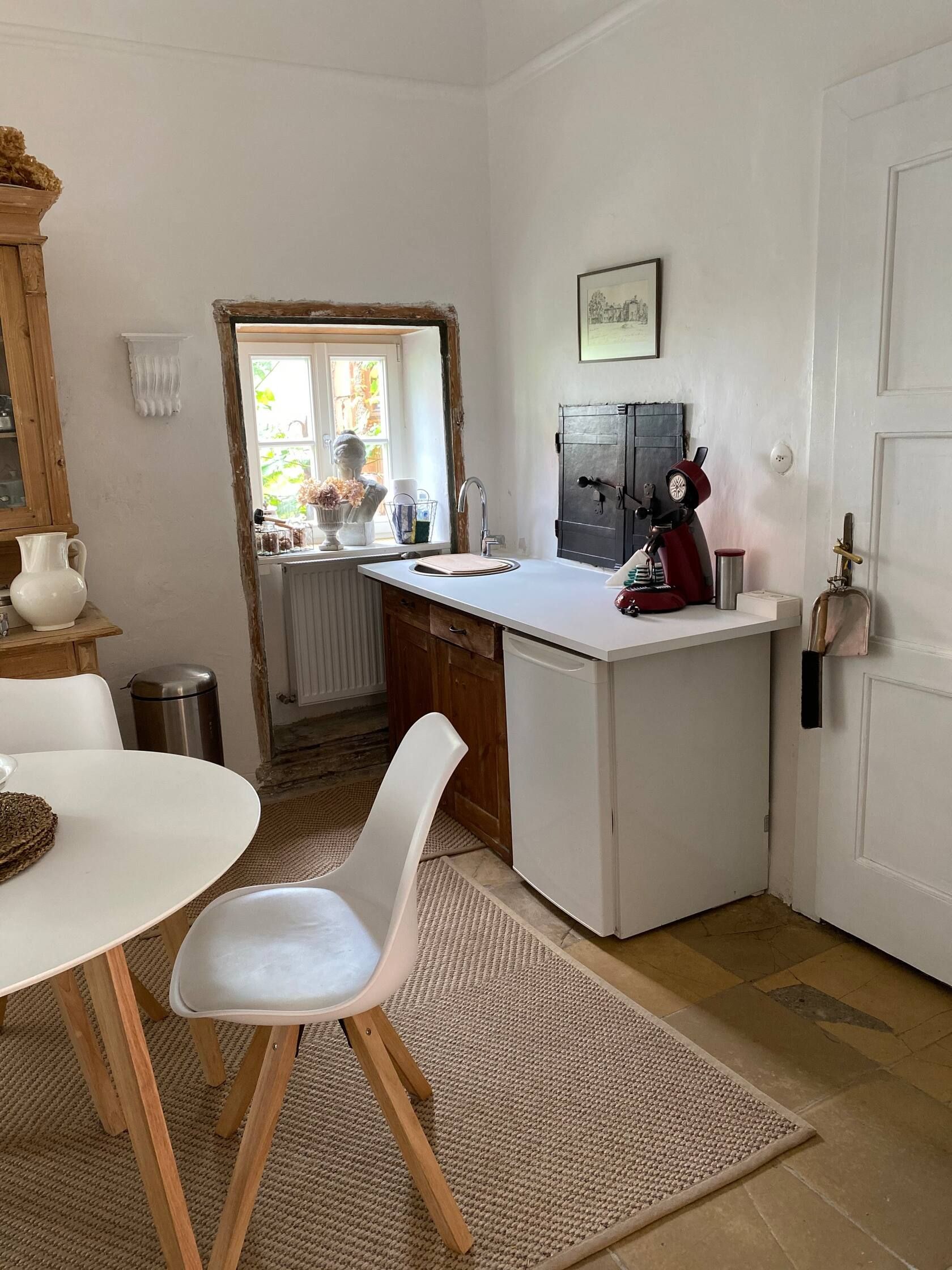 Kuchynský kút so stolíkom, stoličkou, drezom a kávovarom. Okná s dekoráciou a svetlé steny.