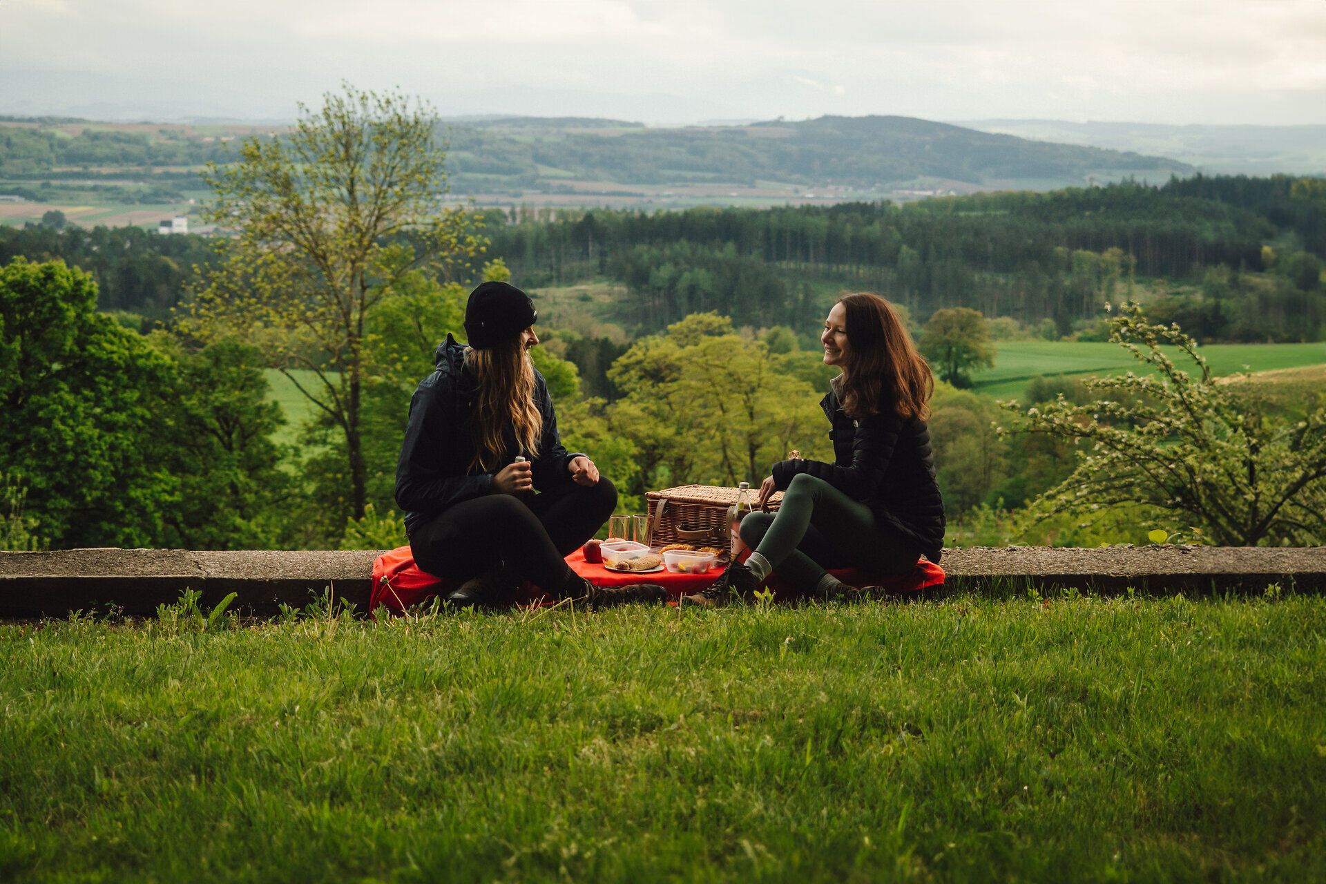 Uprostred malebnej krajiny si dvaja priatelia vychutnávajú pohodový piknik na lúke. Scenéria obklopená sviežou zeleňou a zvlnenými kopcami vyžaruje pokojnú atmosféru, ktorá vás pozýva, aby ste sa zdržali.